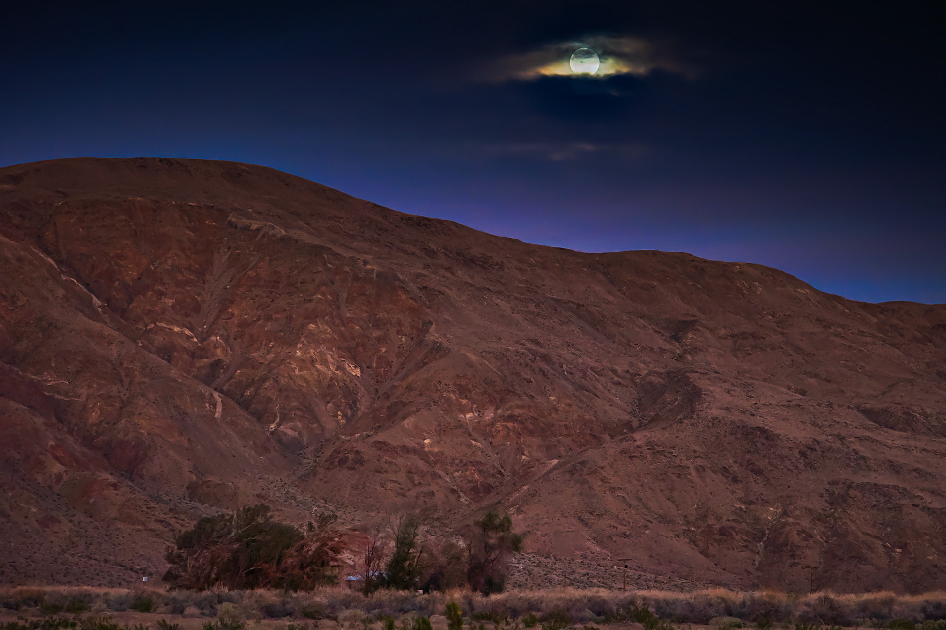 An Eye in the Sky: The rising moon peeks through the cloud cover looking over a farmhouse nestled in a copse of trees at the foot of Coyote Mountain in the Anza-Borrego Desert.