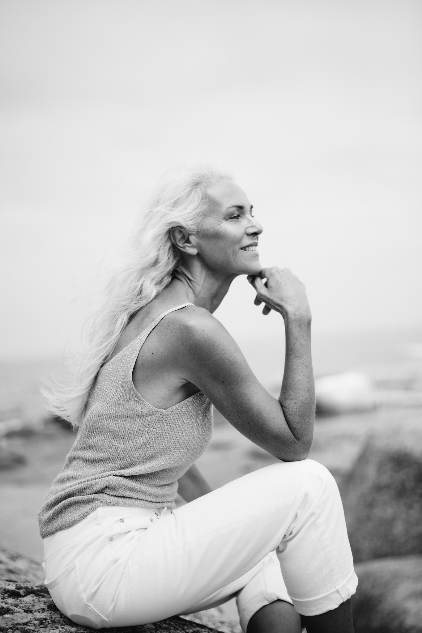 Black and white three-quarter side-profile portrait of woman in white tank top and jeans sitting on rock with knee up, resting chin on hand, looking out, natural candid editorial style, casual women’s outfit inspiration.