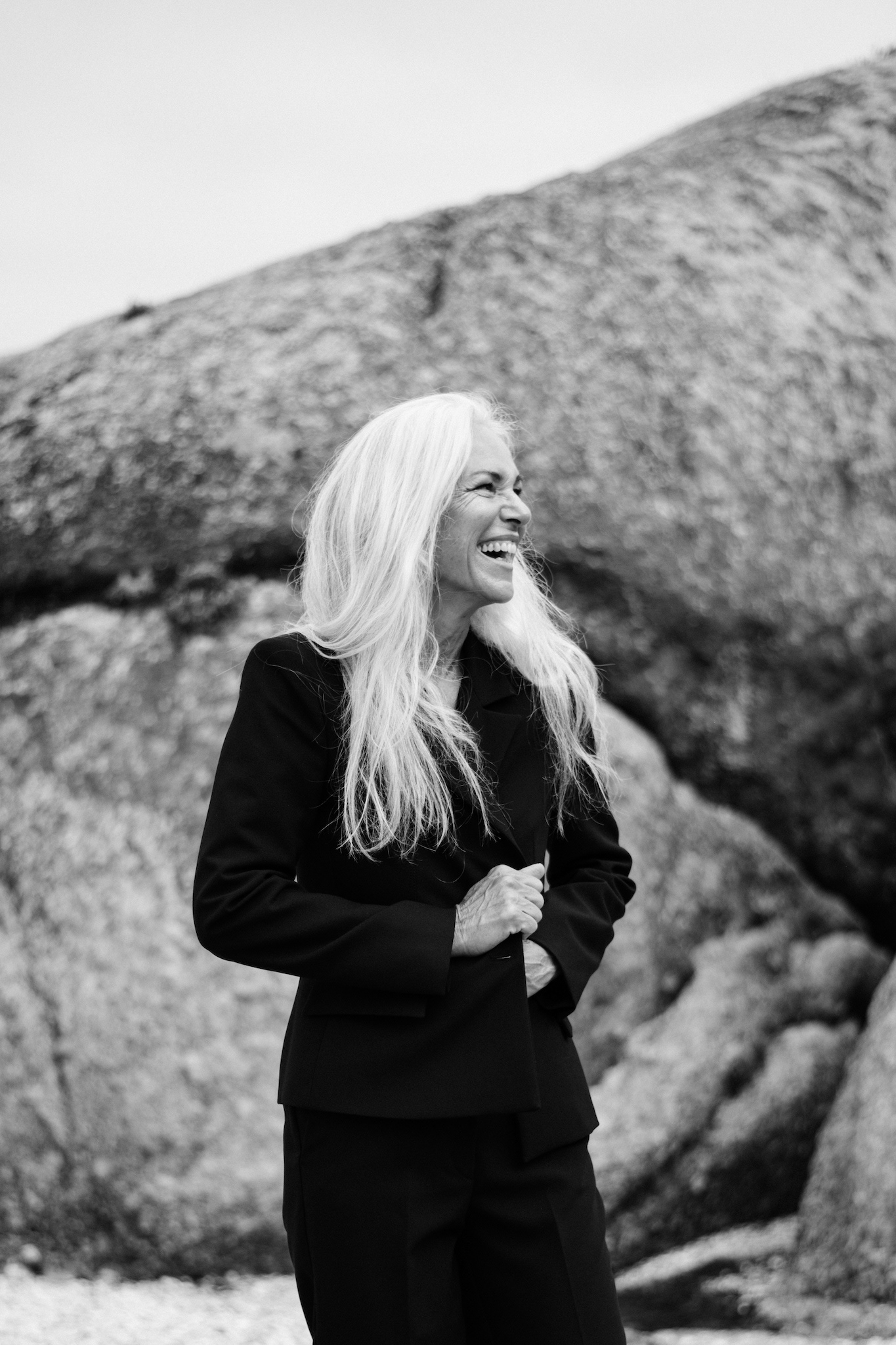 Black and white candid portrait of woman in black suit smiling naturally, adjusting jacket, authentic and joyful editorial moment, storytelling photography capturing real expression.