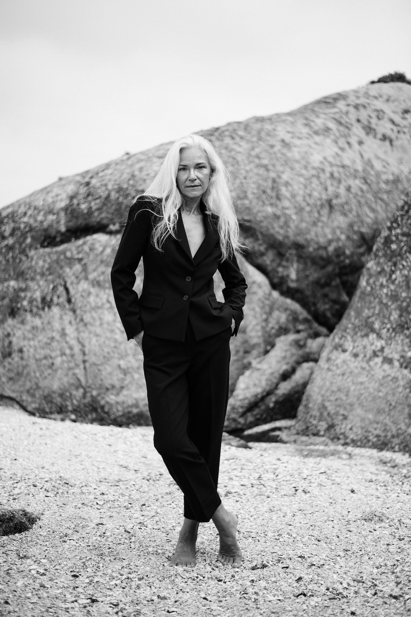 Full-length black and white portrait of woman in black suit standing on Cape Town beach with rock formations, arms in pockets, casual yet strong pose, raw and authentic storytelling portrait.