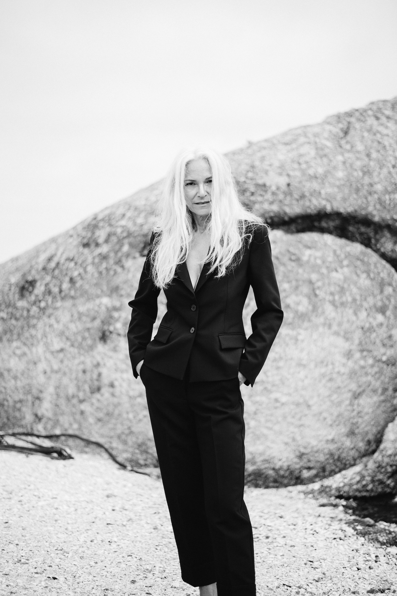 Three-quarter black and white portrait of woman in black suit standing with hands in pockets, looking at camera with subtle expression, raw and authentic editorial style, women’s black suit inspiration.