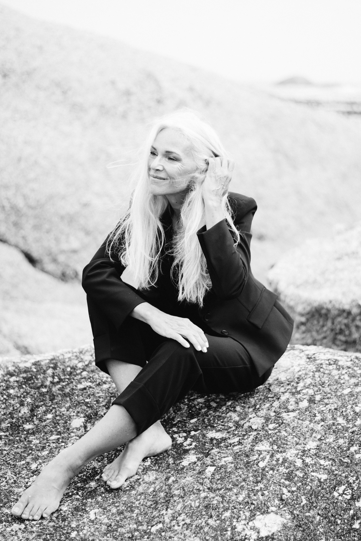 Black and white portrait of woman in black suit sitting on rock, smiling, tucking hair behind ear, candid storytelling moment, editorial and authentic style, women’s fashion inspiration.