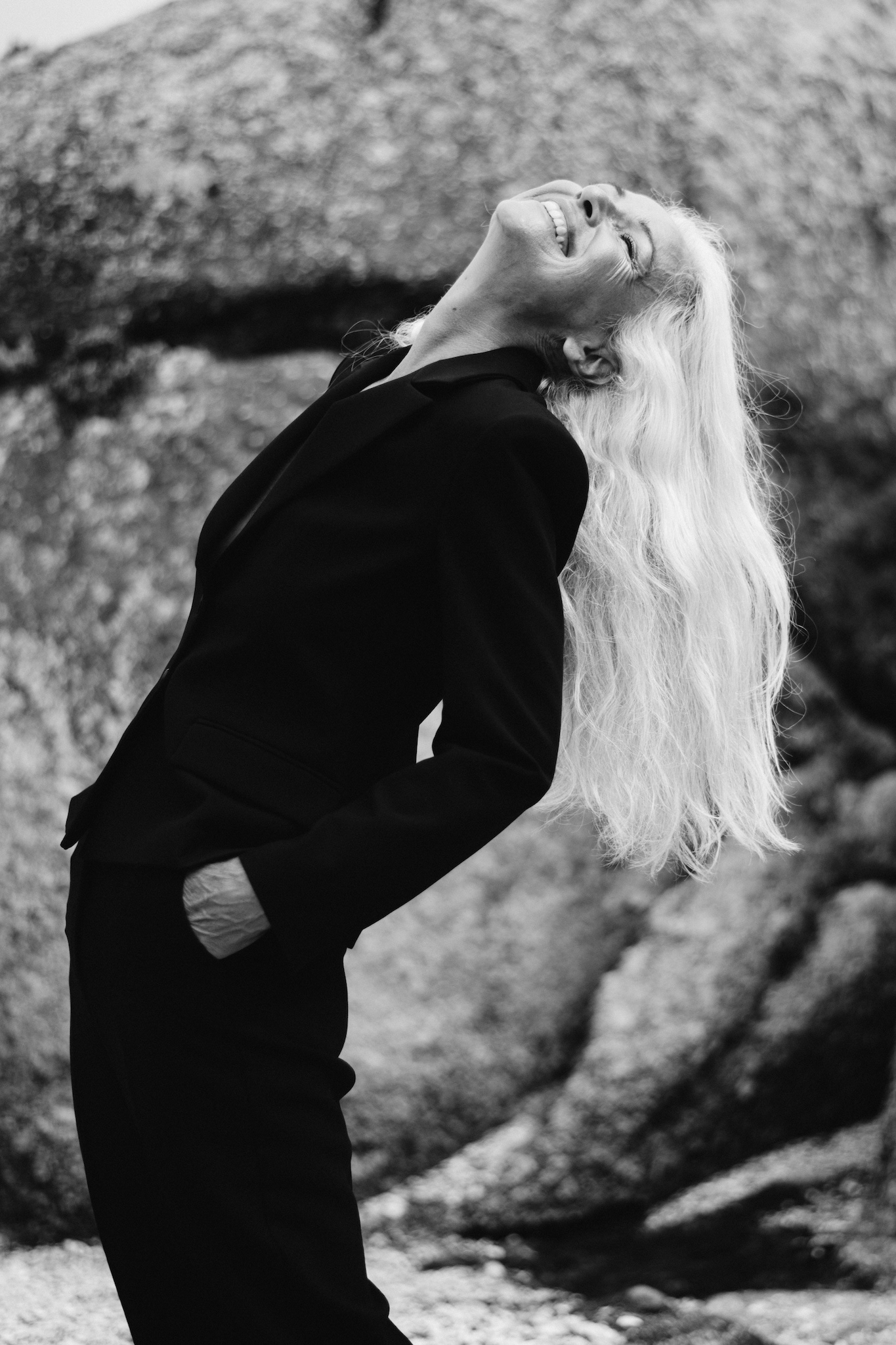 Black and white full-length portrait of woman in black suit laughing while leaning back, hair falling naturally, candid editorial storytelling image, conveying confidence and authenticity.
