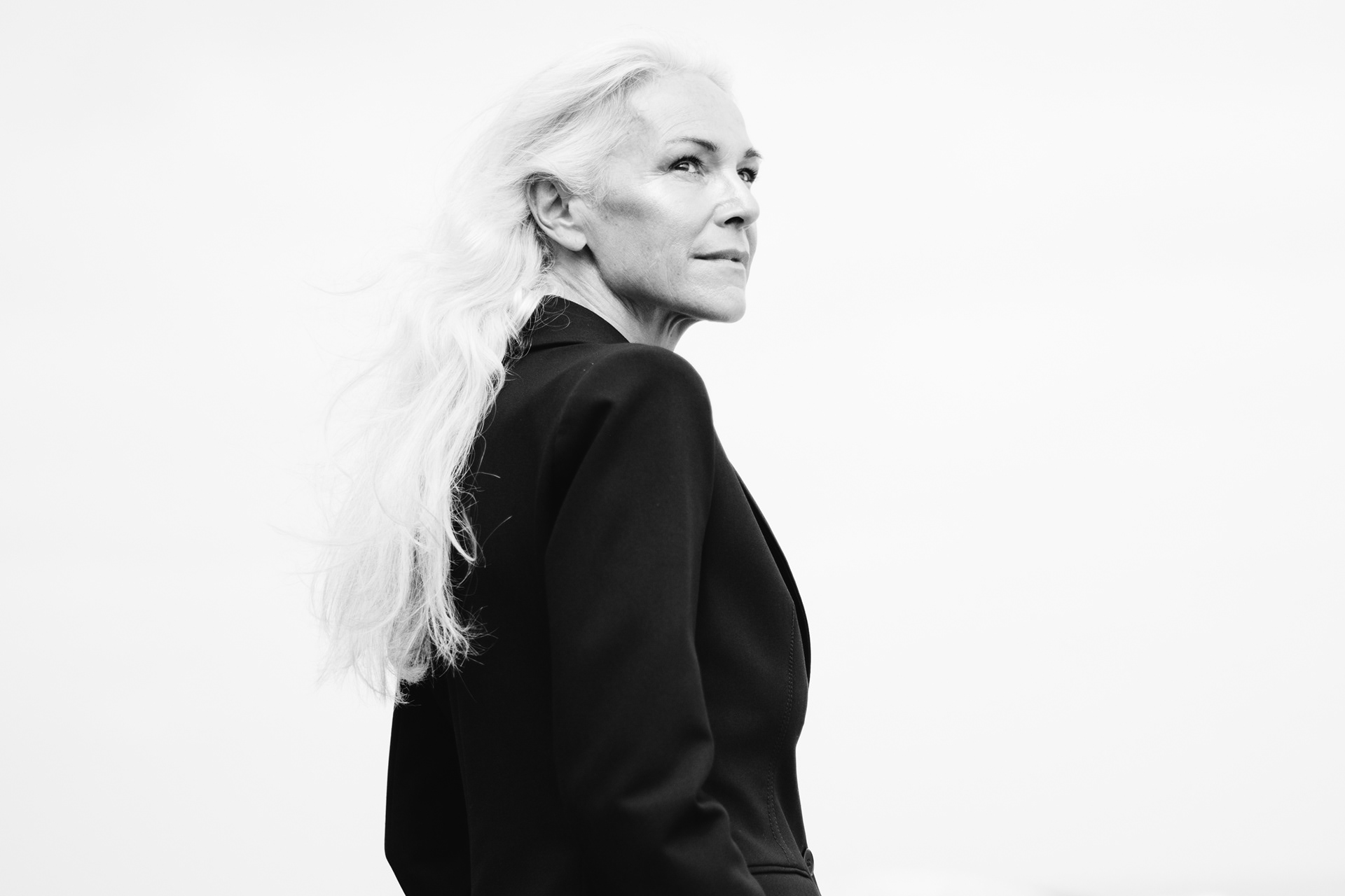 Black and white landscape portrait of woman in black suit on sandy beach, hair blowing in the wind, gazing away from camera, authentic editorial photography capturing natural beauty.