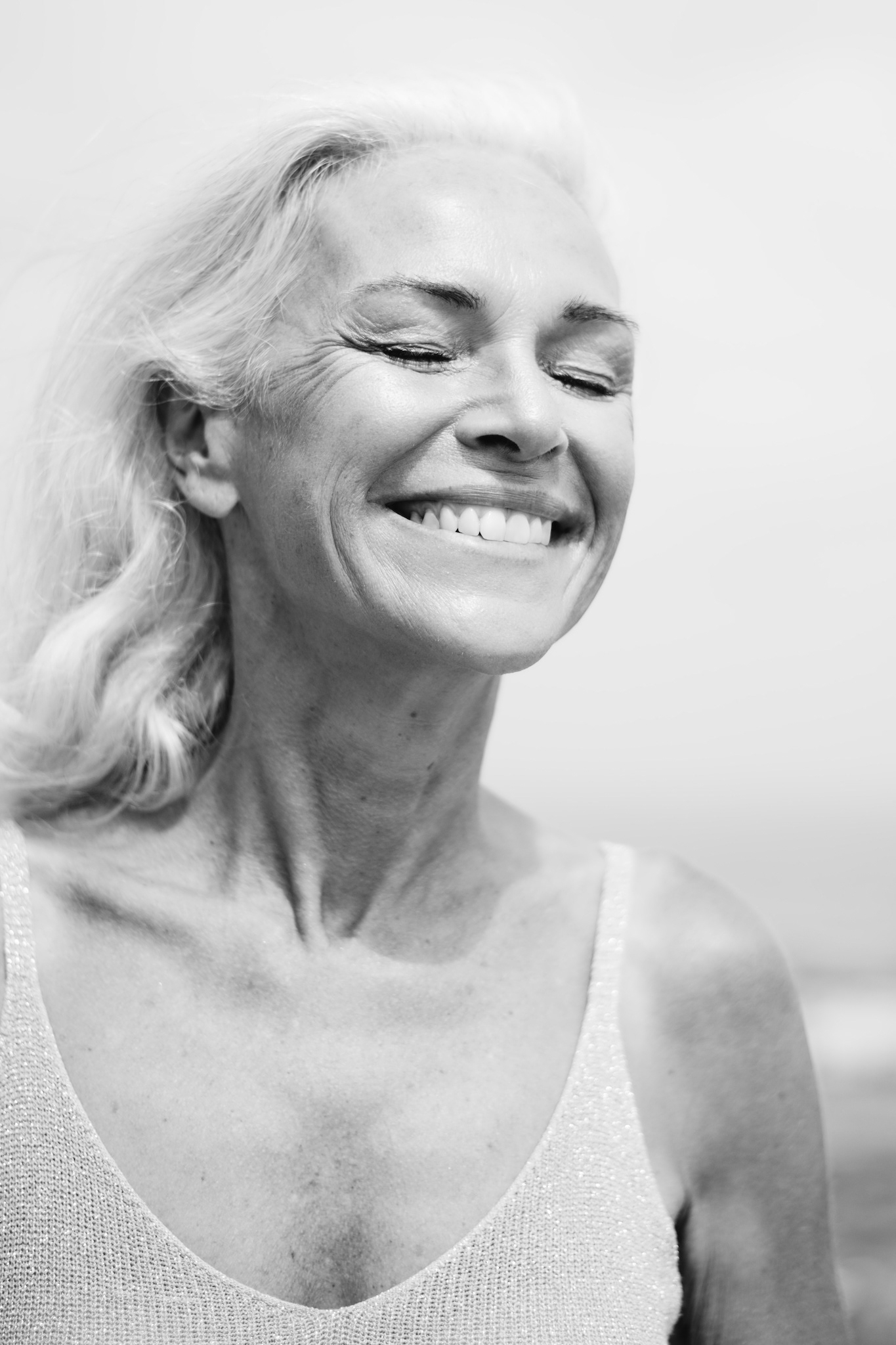 Close-up black and white portrait of woman in white tank top and jeans, smiling with eyes closed, candid editorial storytelling moment, authentic and joyful photography, casual outfit inspiration for women.