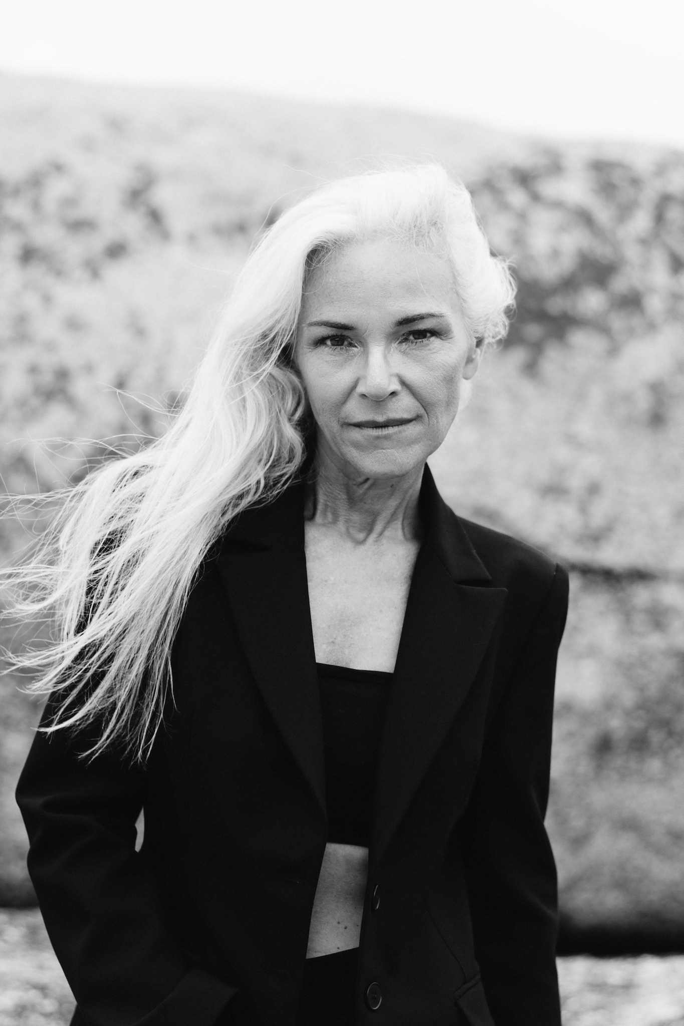 Black and white close-up portrait of woman in black suit with hair blowing to the side, subtle reveal of tube top underneath jacket, strong editorial and storytelling portrait, confidence-focused.