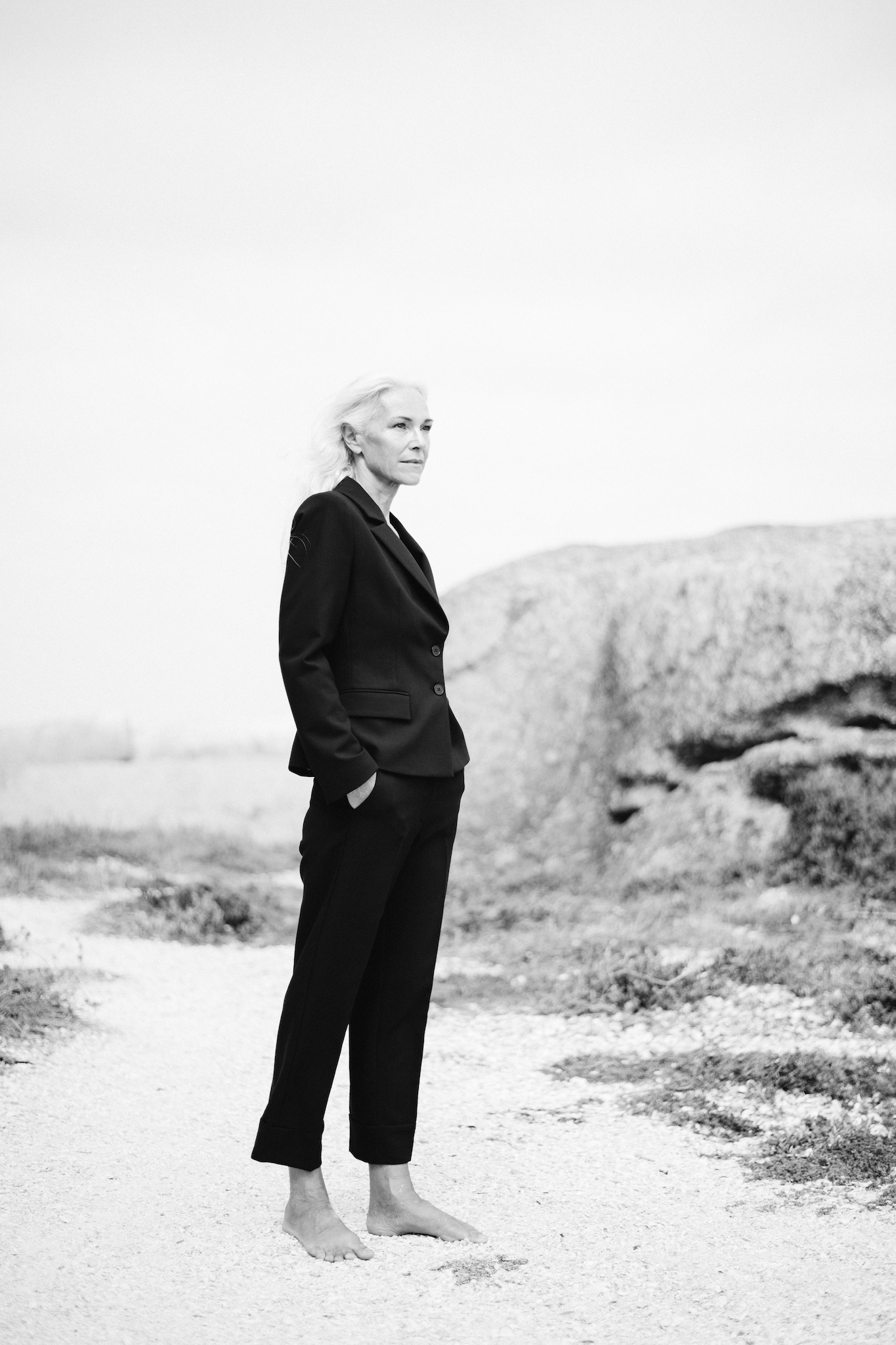 Full-length black and white portrait of woman in black suit standing slightly diagonal to camera, looking off to the side, natural editorial composition, storytelling and authentic style.
