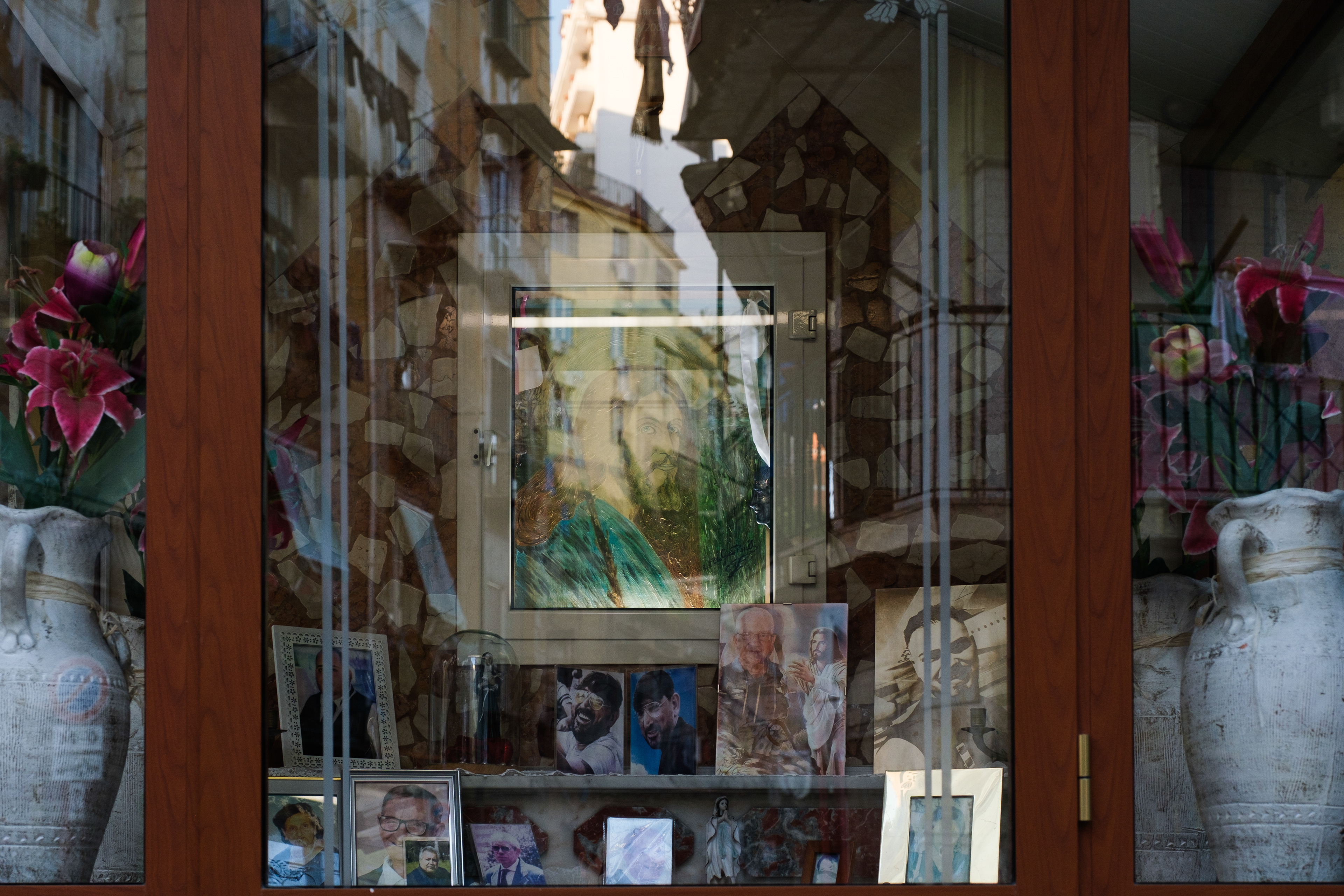 Naples (Italy): A street-facing window display houses a memorial altar with framed portraits of the deceased in the Rione Sanità district, with the glass reflection visually merging the interior shrine with the surrounding city architecture.