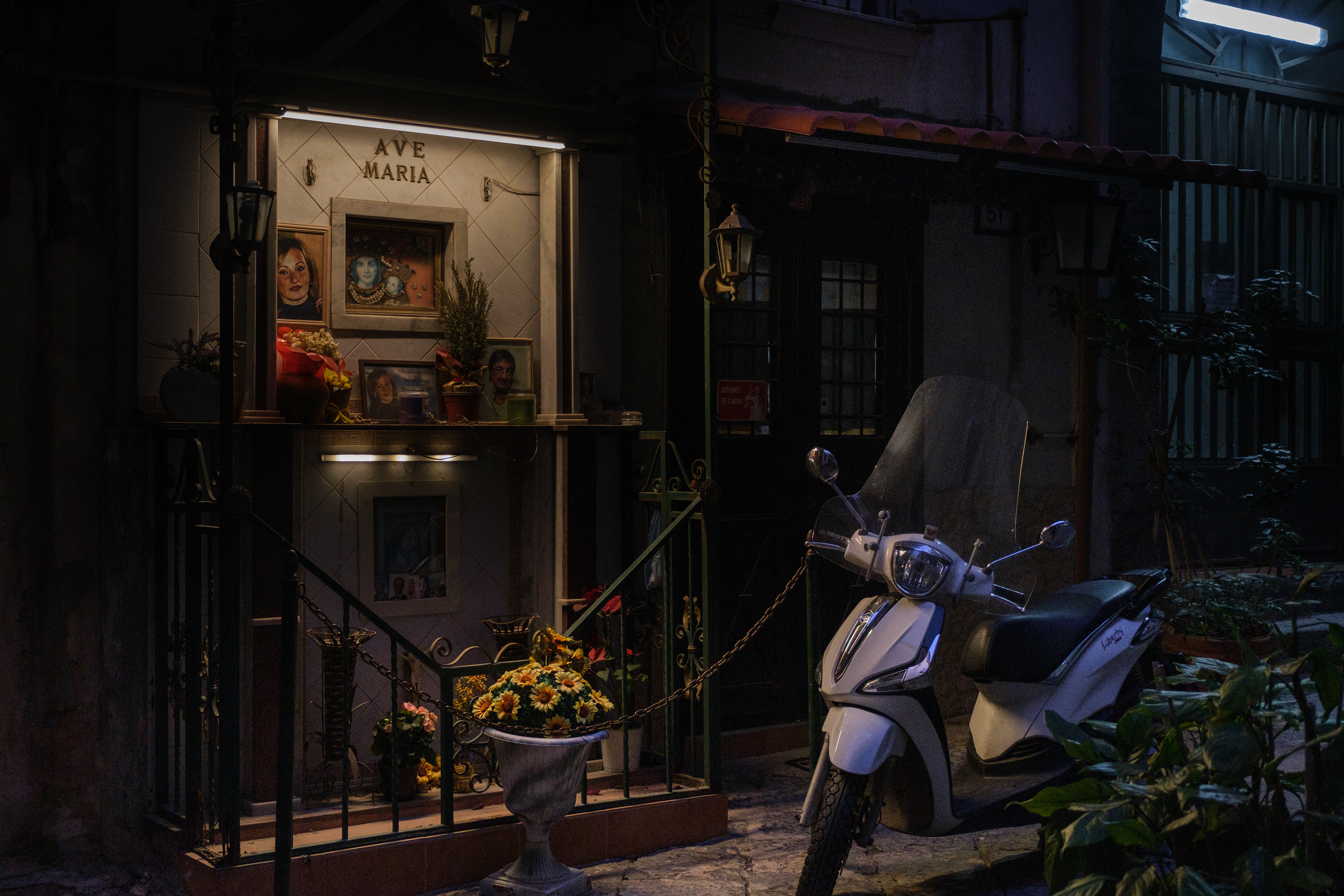 Naples (Italy): A street shrine inscribed with "A VE MARIA" is illuminated at night in the Rione Sanità district, featuring framed portraits of the deceased and floral arrangements next to a parked motor scooter.