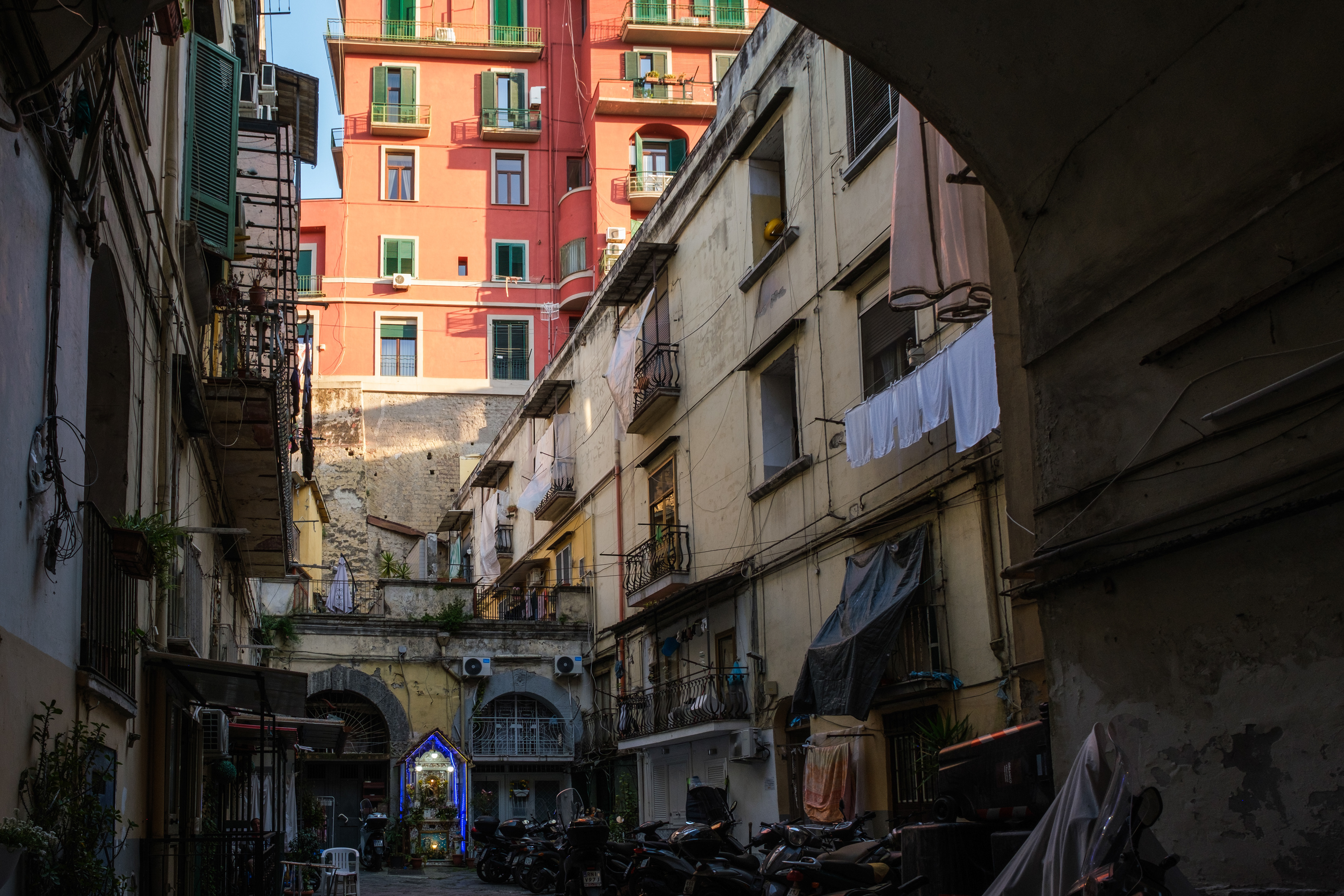Naples (Italy): A view of a narrow residential courtyard in the Rione Sanità district, featuring hanging laundry, parked motor scooters, and a brightly lit blue neon edicola votiva (street shrine) at the far end.