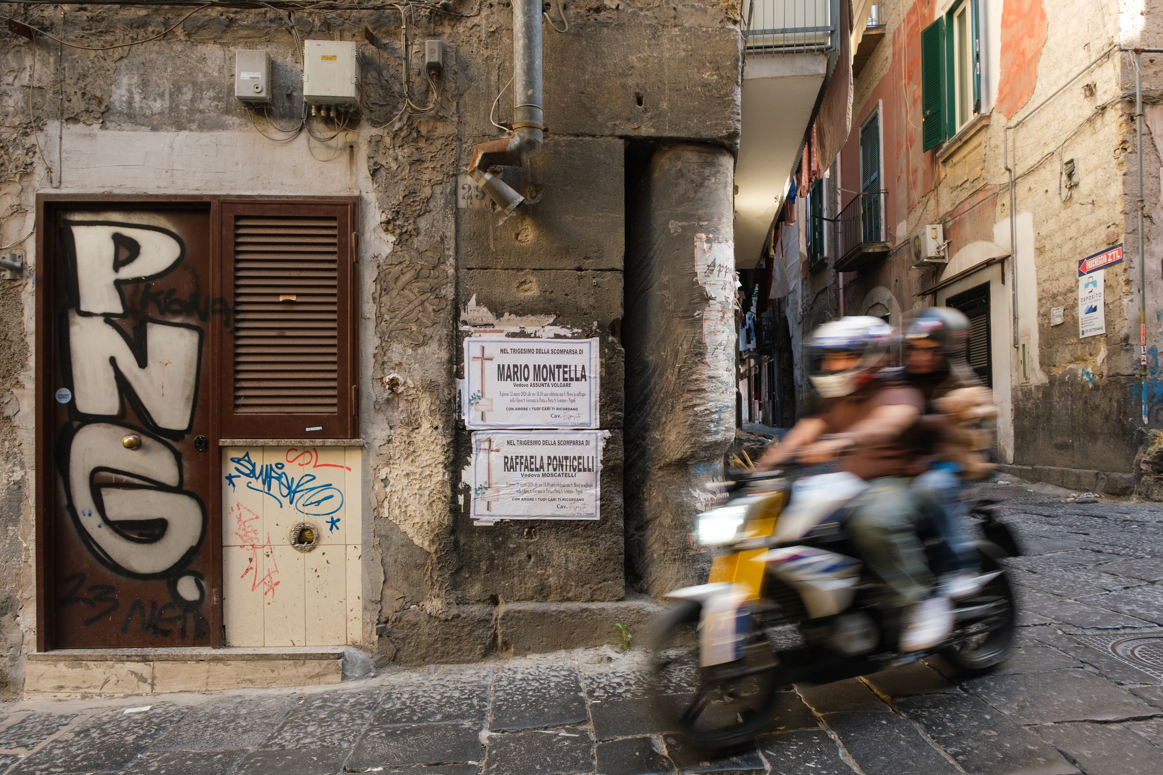 Naples (Italy): A motorcycle passes in motion blur next to a weathered stone wall displaying death notice posters in a narrow street of the Rione Sanità district.