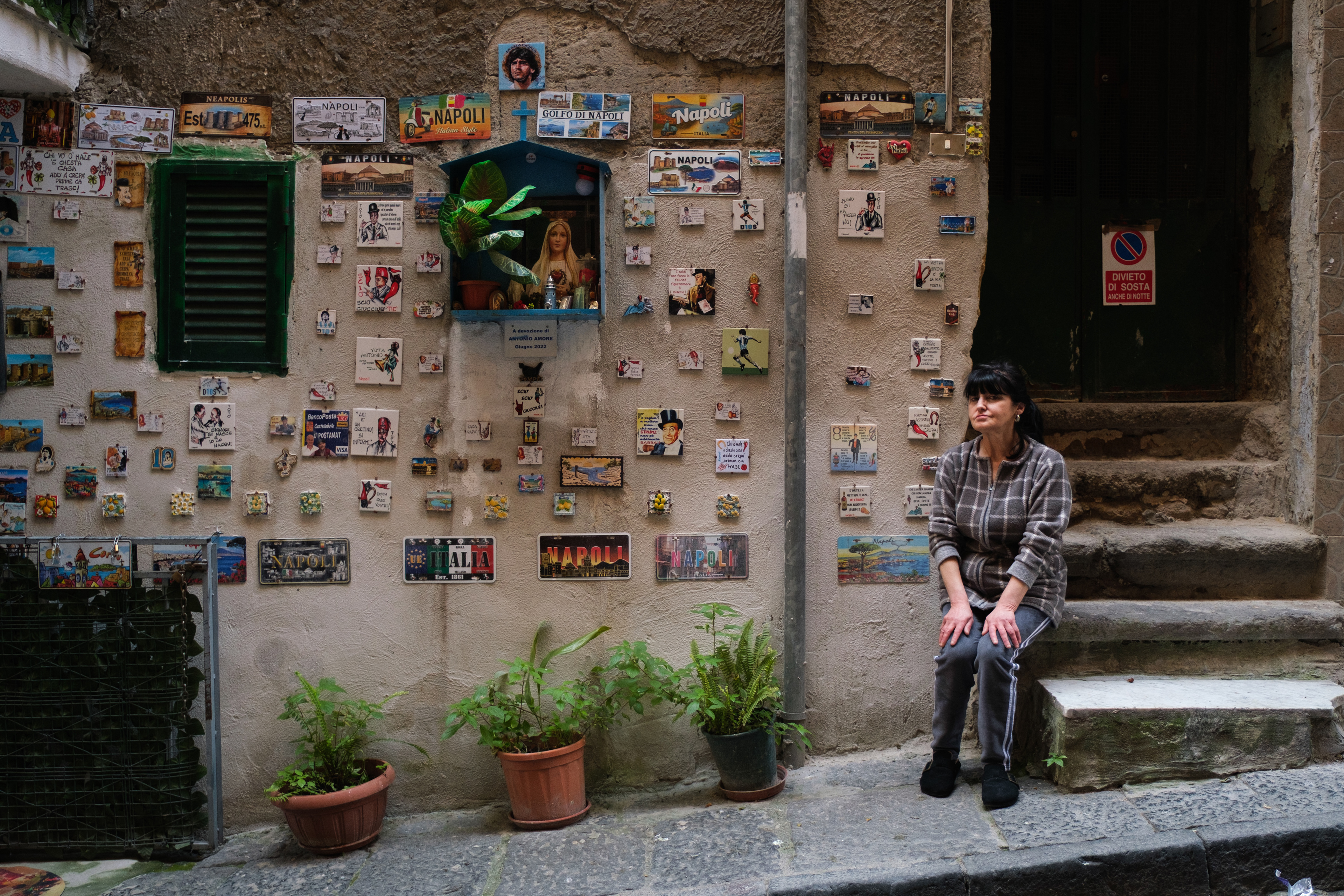 Naples (Italy): Nuncia Amore sits on stone steps next to a street shrine built in memory of her late brother, Antonio Amore, in the Rione Sanità district. The wall features a decorative mural of tiles assembled by Antonio during his lifetime.