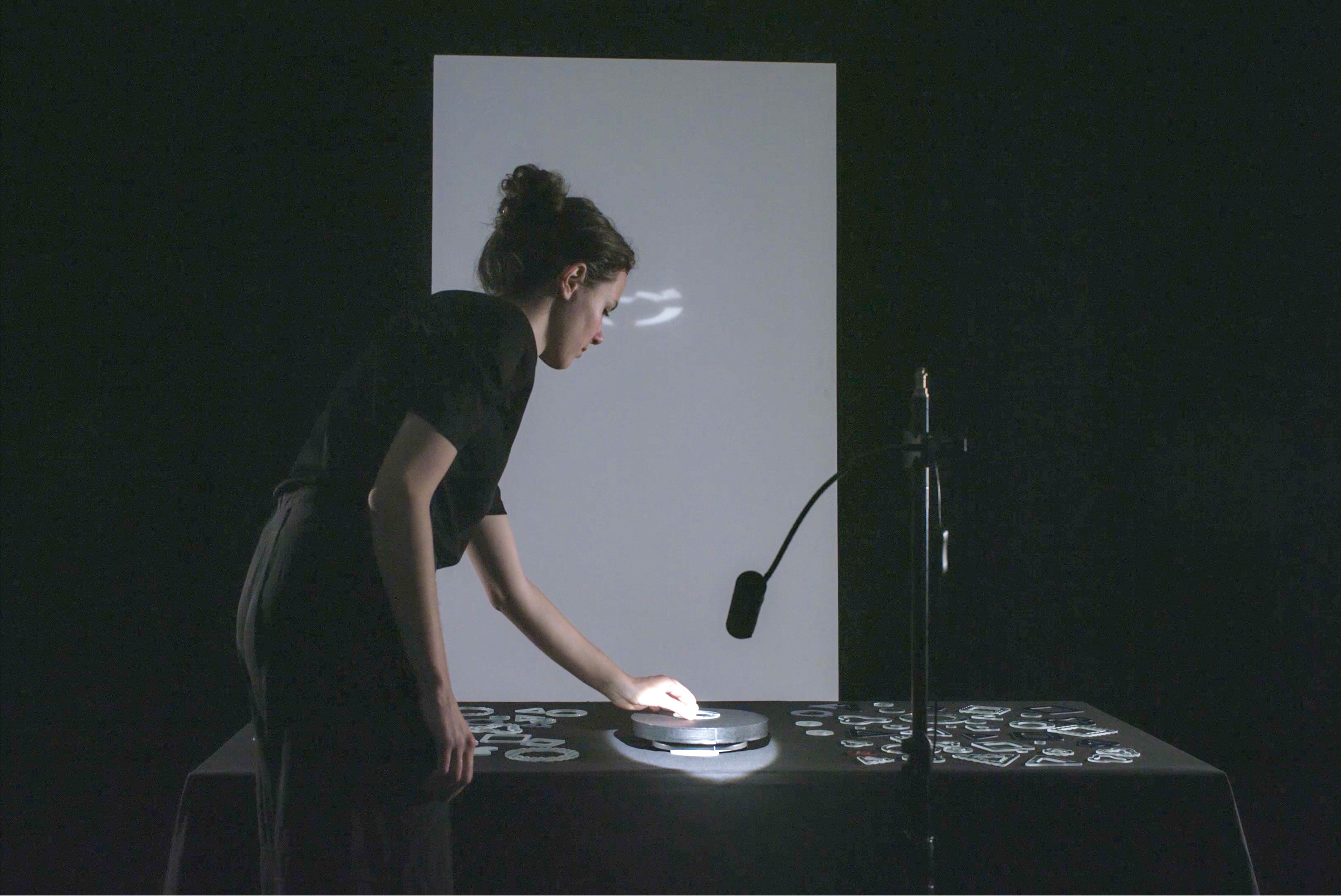 In a dark room, a person in black leans over a table covered with black cloth and places a glass shape on a round lit platform, while light from it appears on a tall white screen behind. More glass shapes are spread across the table, and a black cylinder on a gooseneck stand stands at the right.