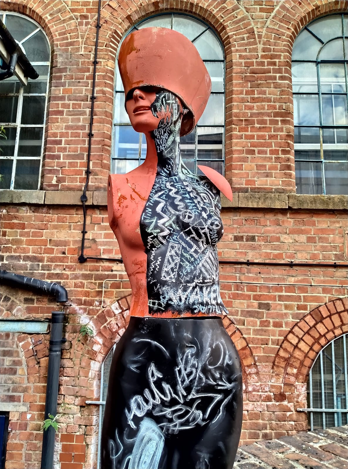 Three photographs of a distorted human mannequin inscribed with chalk. The figure has no arms, and its eyes are obscured by a helmet. The body is bisected, with the helmet, shoulder pad and half the torso in red. Starting with defiantly monstrous fangs painted on half of the jaw, other areas are covered in black paint and have been filled with chalked text. The text on the top half combines Green Chinese characters, blue English words and white sigils. The upper leg piece is painted all black, with a large white sigil and a faded image of a cassette tape. The sculpture is set in front of the BACKLIT gallery building: an old redbrick factory with large arched windows under a blue sky with white clouds. The angles the figure is shot in mirror its geometry. There is a high visual contrast, as if viewed in an altered state: everything is vivid even when it is indecipherable.