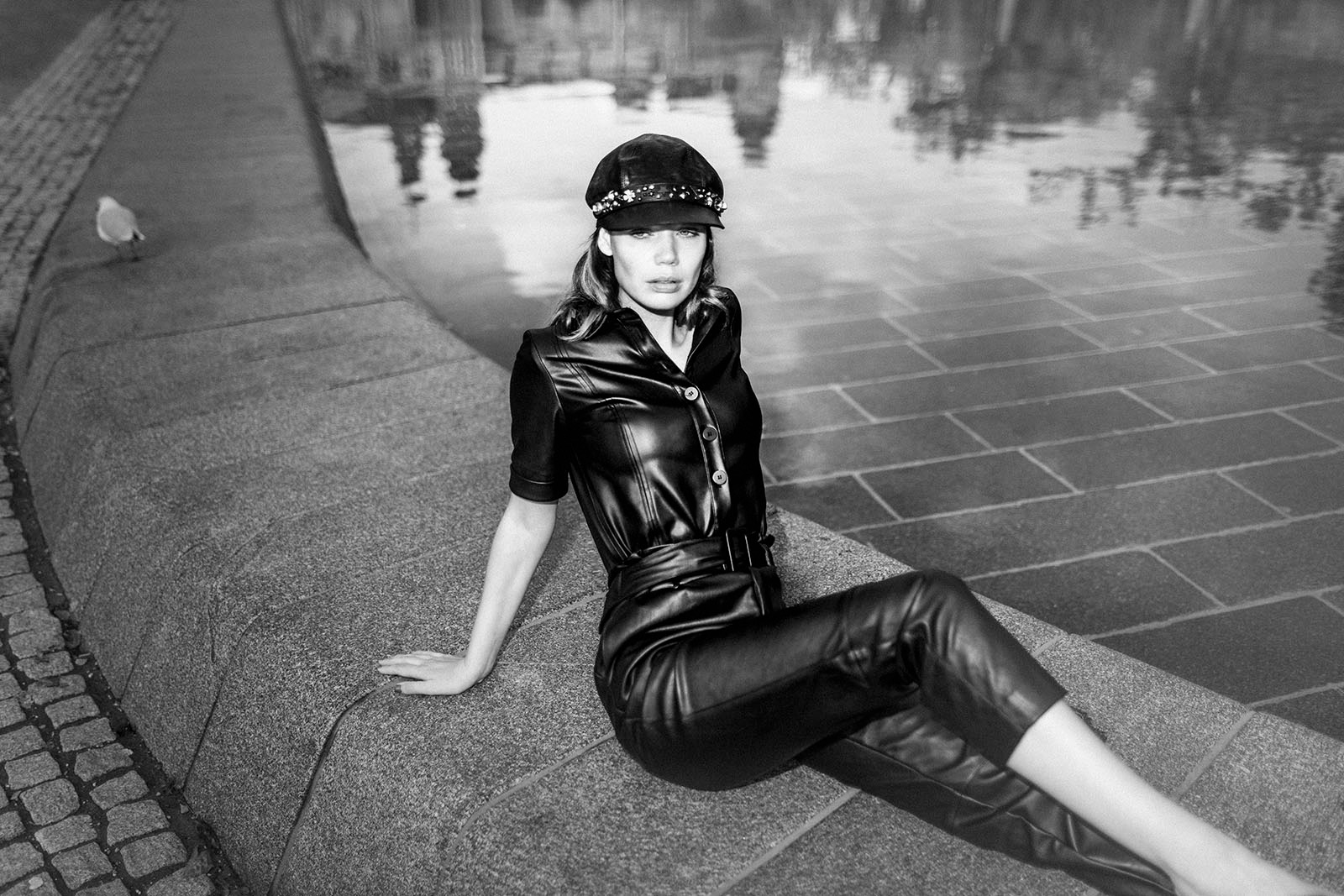 Zwartwit foto van model in zwart leer latex met pet op zittend naast vijver in Amsterdam
