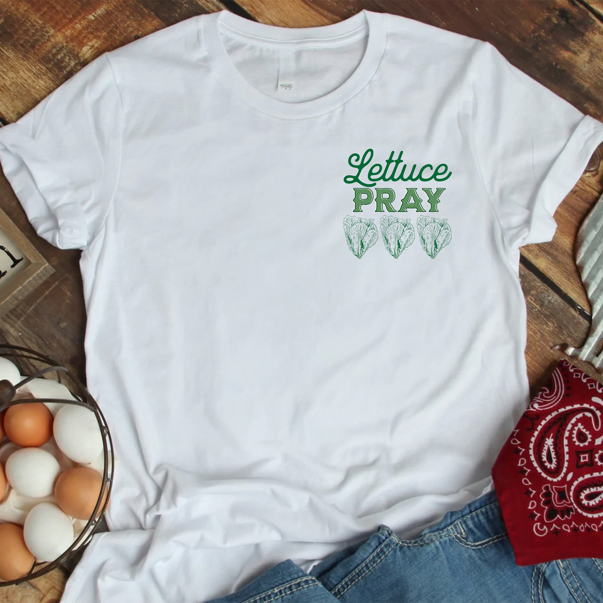 White t-shirt that has romaine lettuce & the words Lettuce Pray, laying on hardwood floor surrounded by a basket of fresh chicken eggs and a red bandana