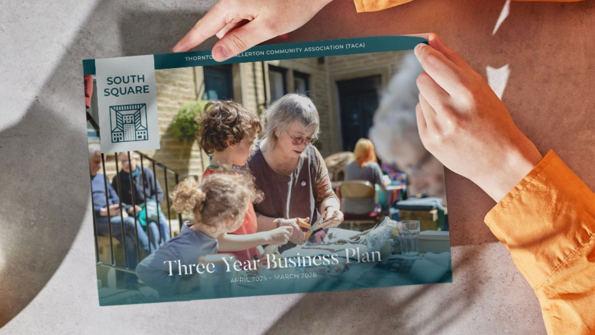 Cover for South Square Centre three year business plan. Features an older woman cutting up some paper alongside two children. Person with orange shirt sleeves holds the booklet.