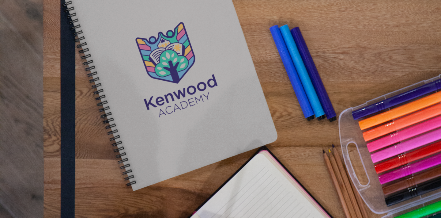 close up of kenwood academy logo on a notebook alongside multi coloured pens on a wooden table