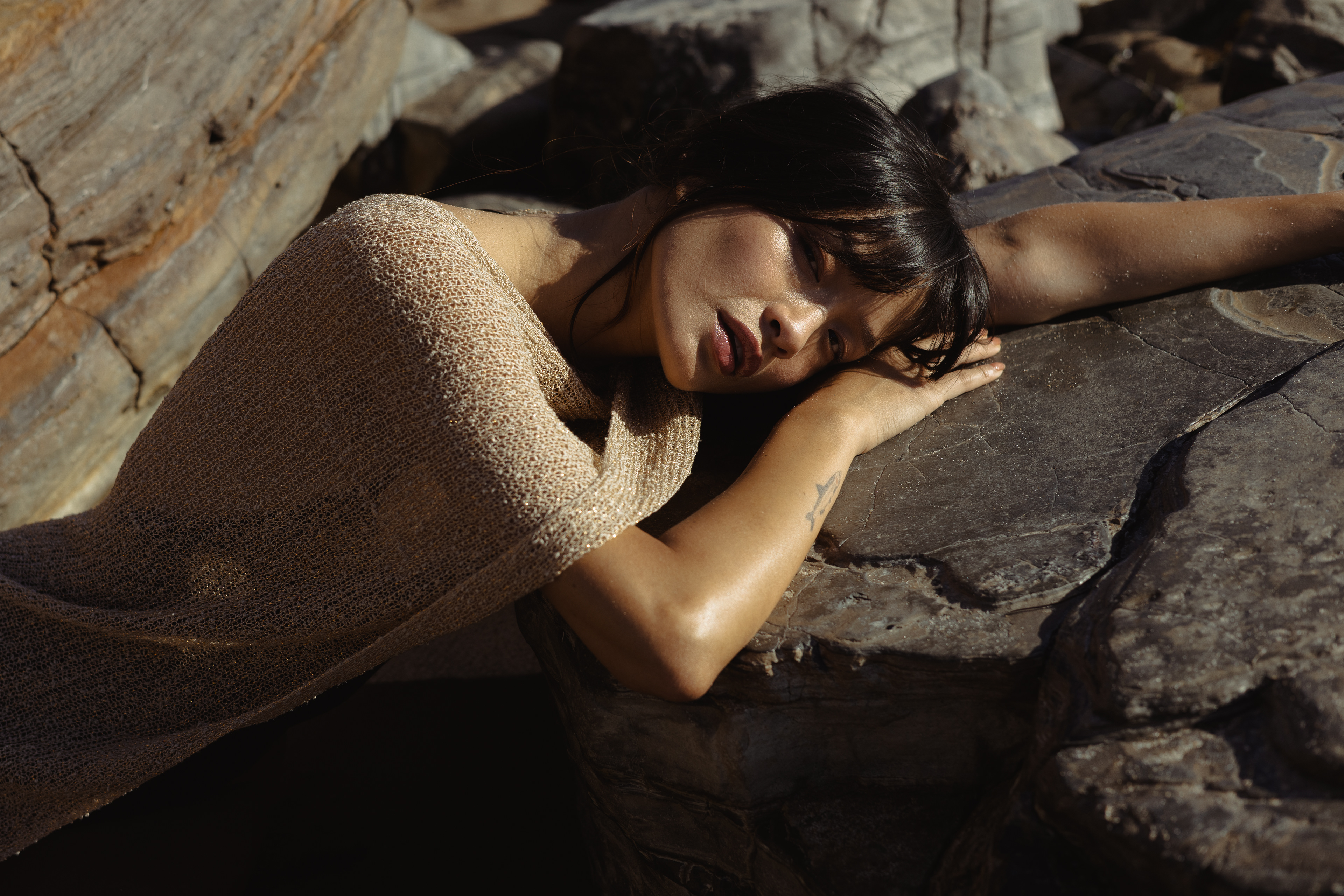 Editorial fashion portrait of a woman in a textured beige knit top resting her head on her arms against sunlit coastal rocks.