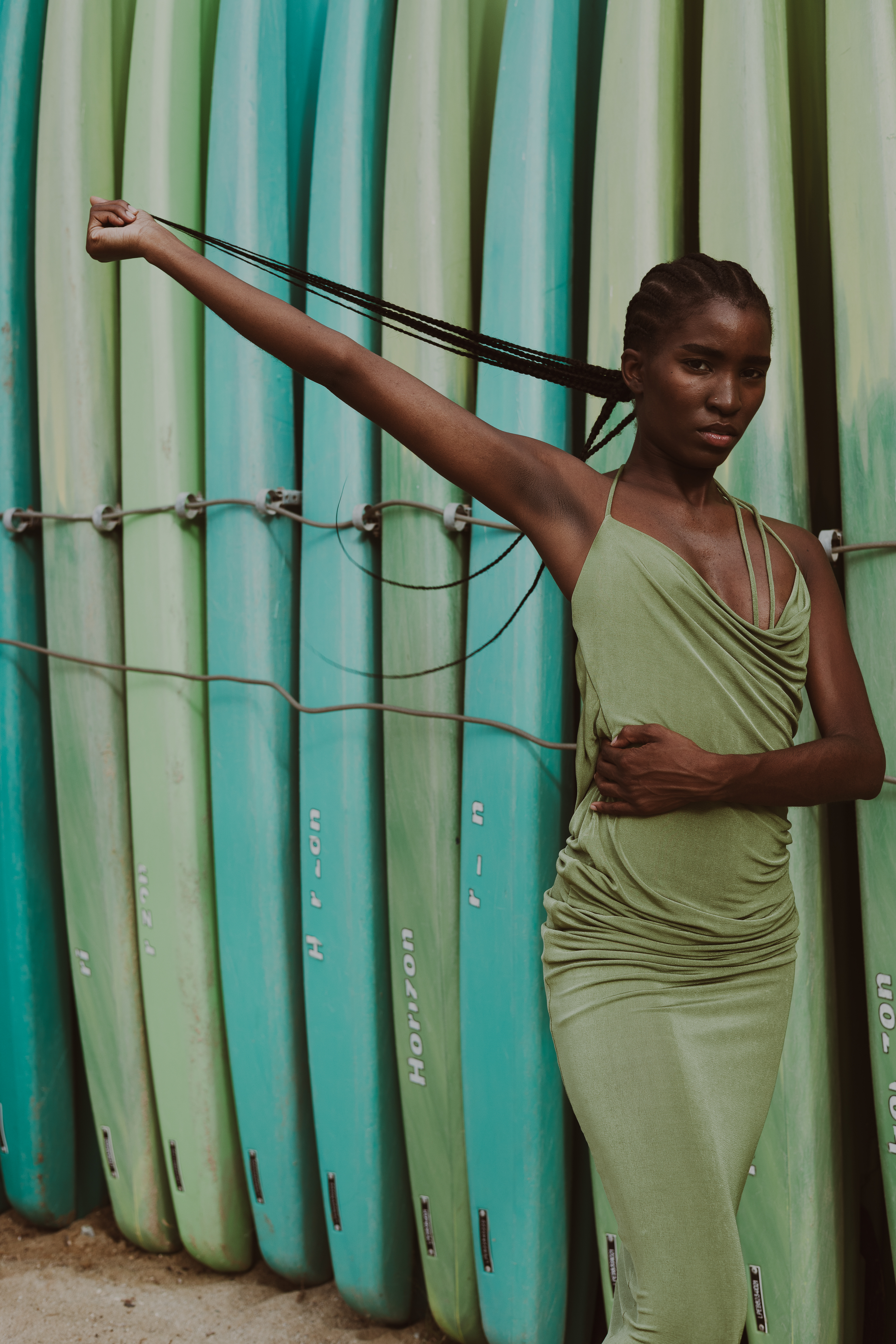 Editorial fashion portrait of a woman in a draped sage-green dress, extending one arm while holding her long braids against a backdrop of turquoise and green surfboards.