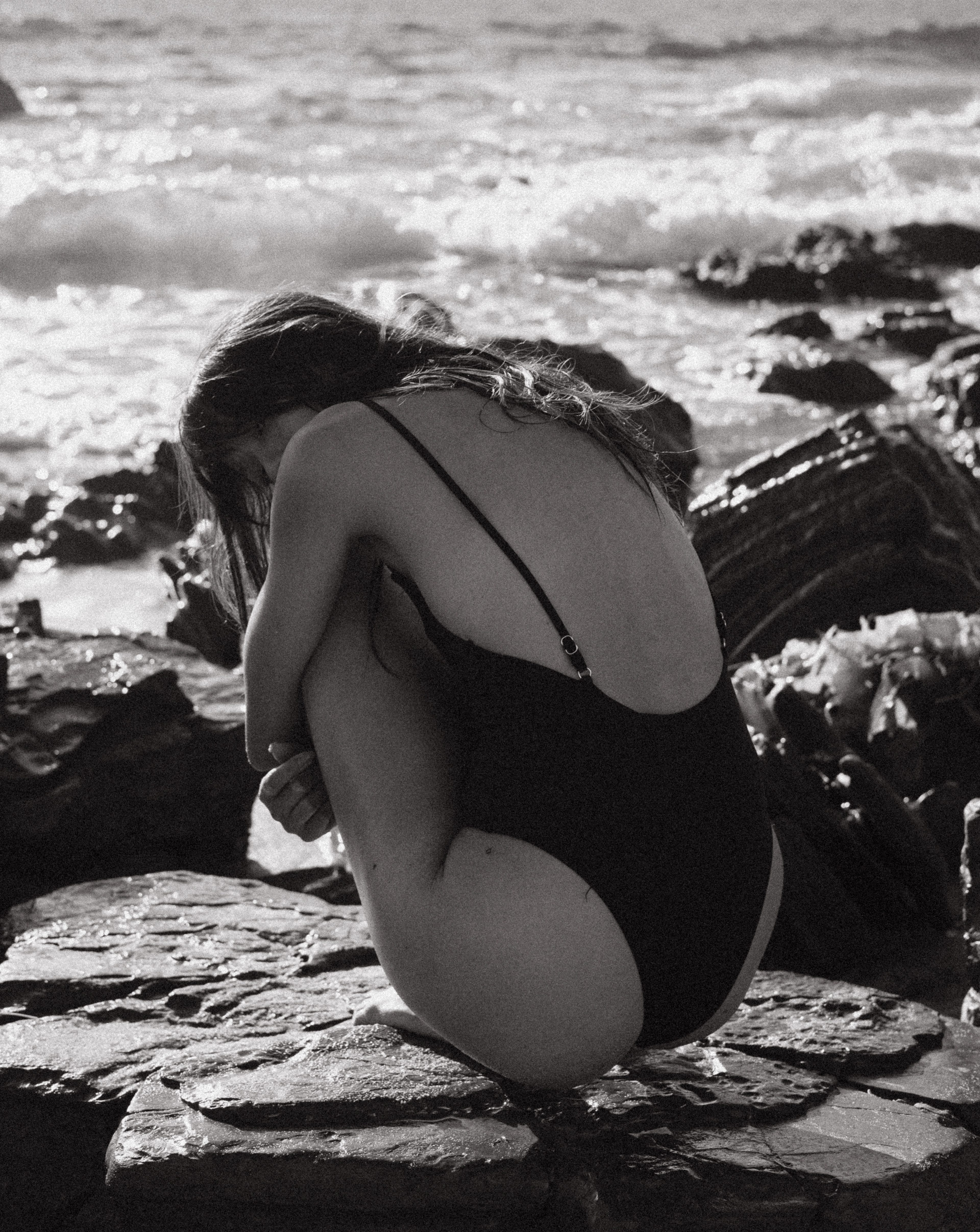 Black-and-white fashion photograph of a woman in a black bodysuit curled up on coastal rocks, with ocean waves in the background.