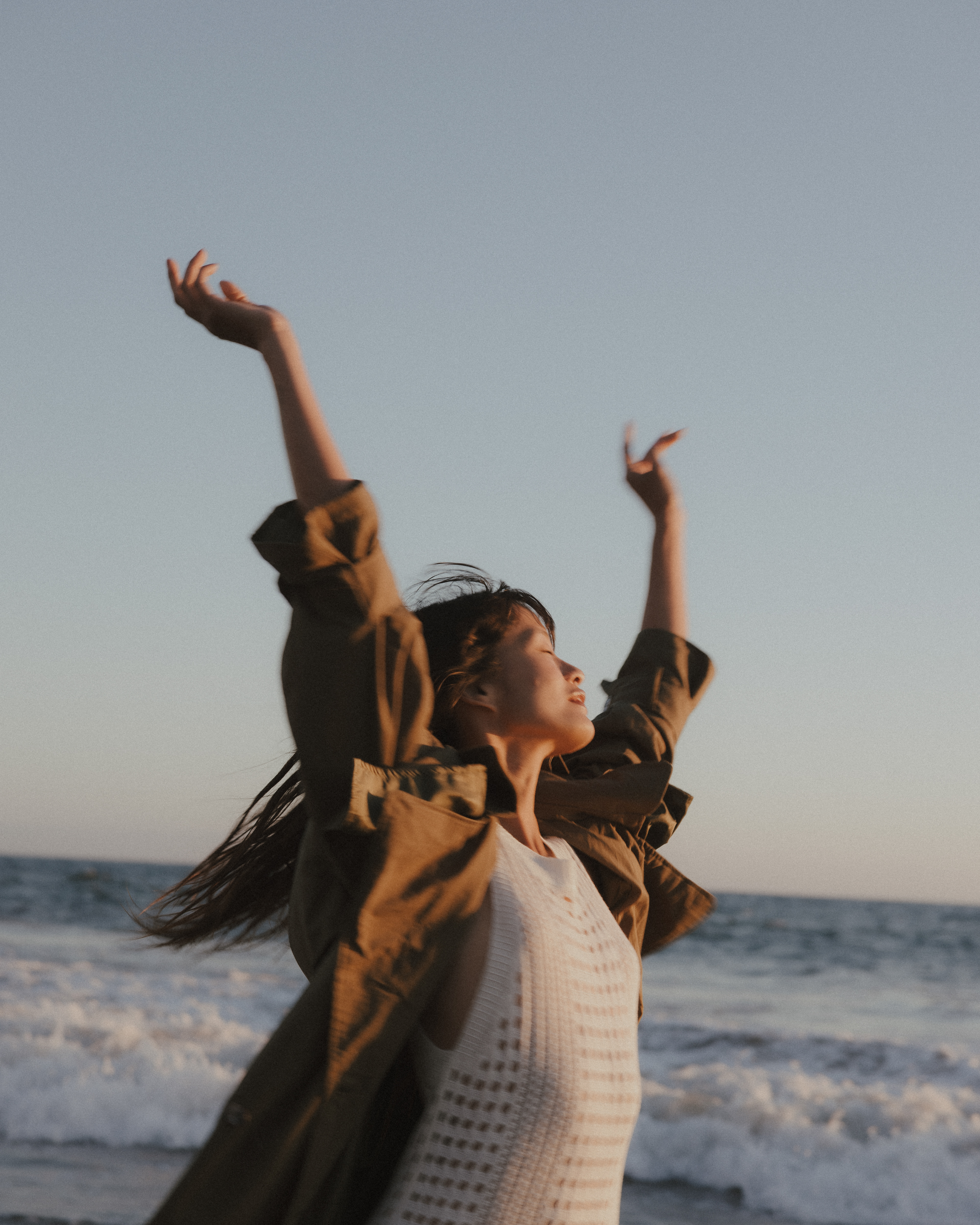 Fashion editorial photograph of a woman wearing an olive-green jacket over a white knit dress, raising both arms against the ocean sky.