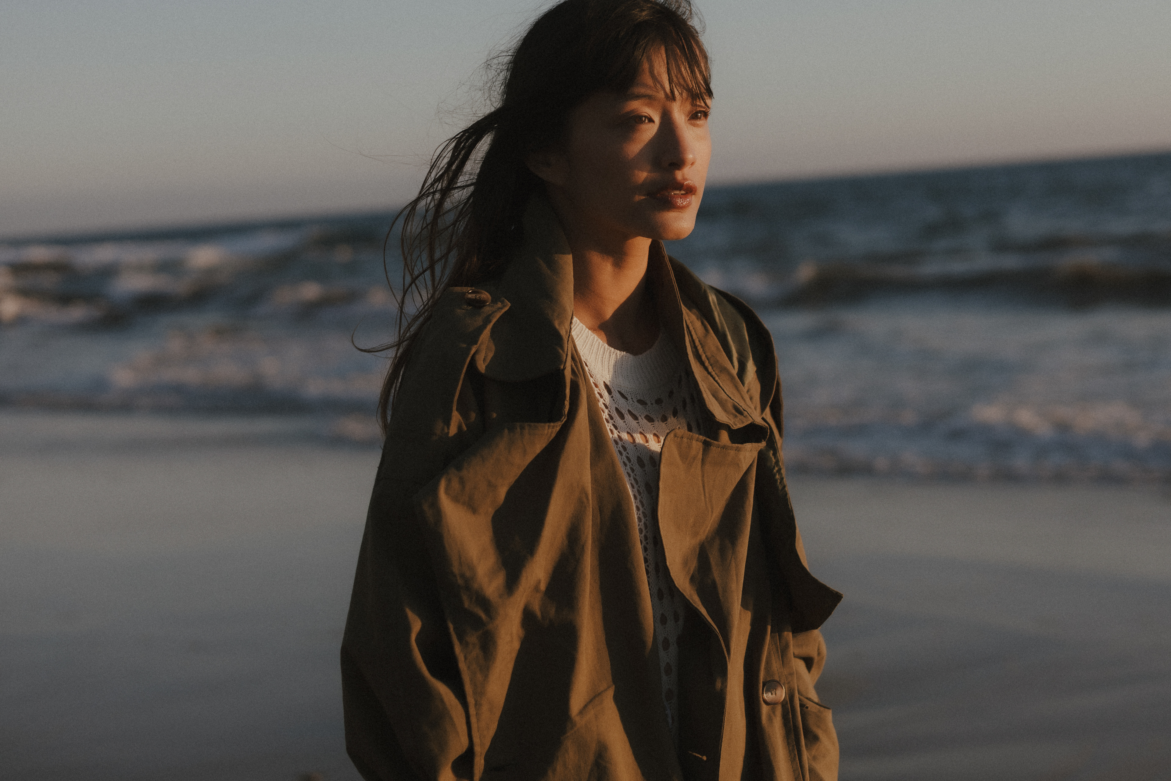 Editorial fashion portrait of a woman in an oversized olive-green jacket and white knit dress standing on the beach in warm golden-hour light.