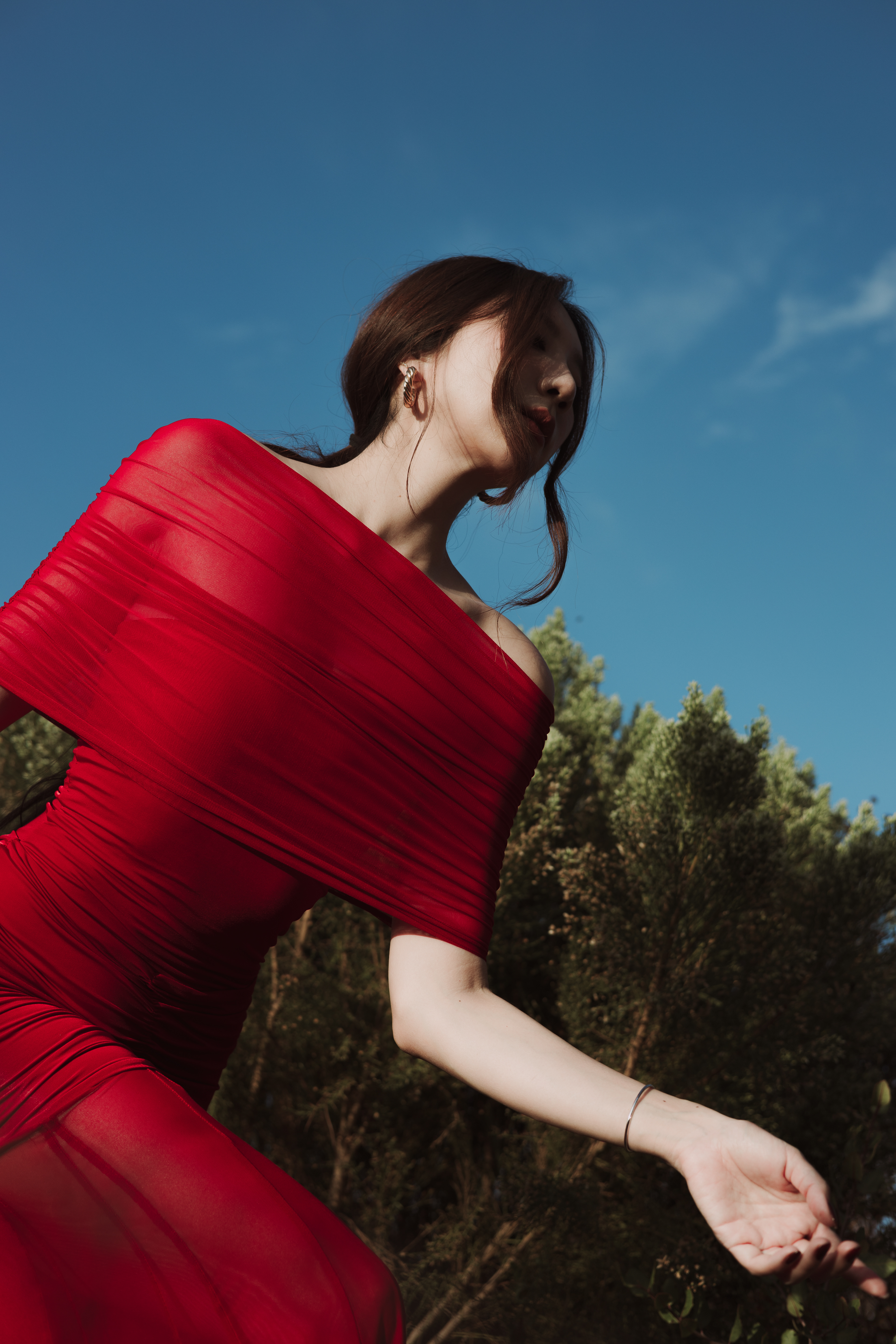 Editorial fashion portrait of a woman wearing a vibrant red off-the-shoulder draped dress, captured in a dynamic pose against a clear blue sky and natural landscape.