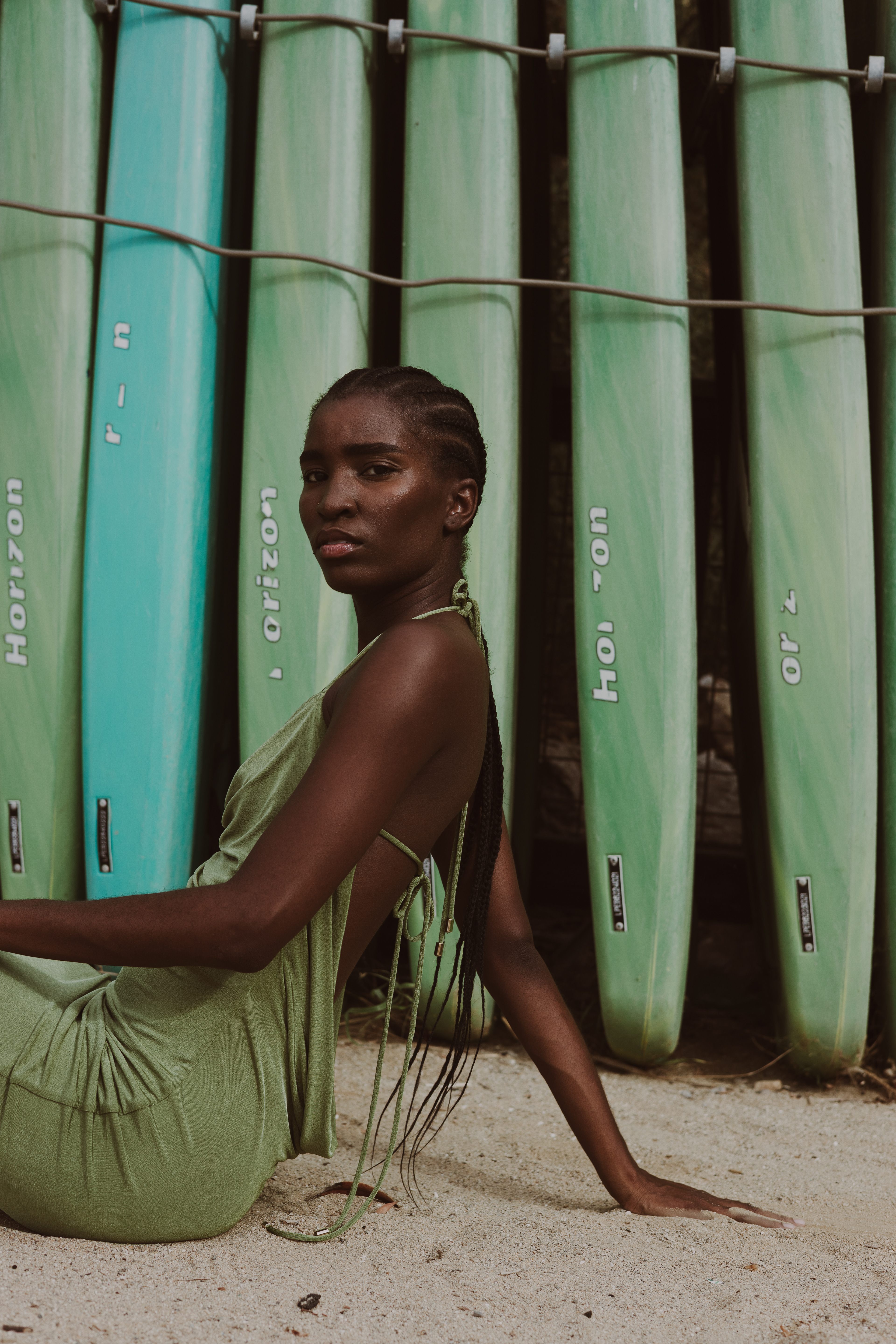 Fashion editorial portrait of a woman in a sage-green dress seated on the sand, leaning back on one arm with long braids against a backdrop of green surfboards.