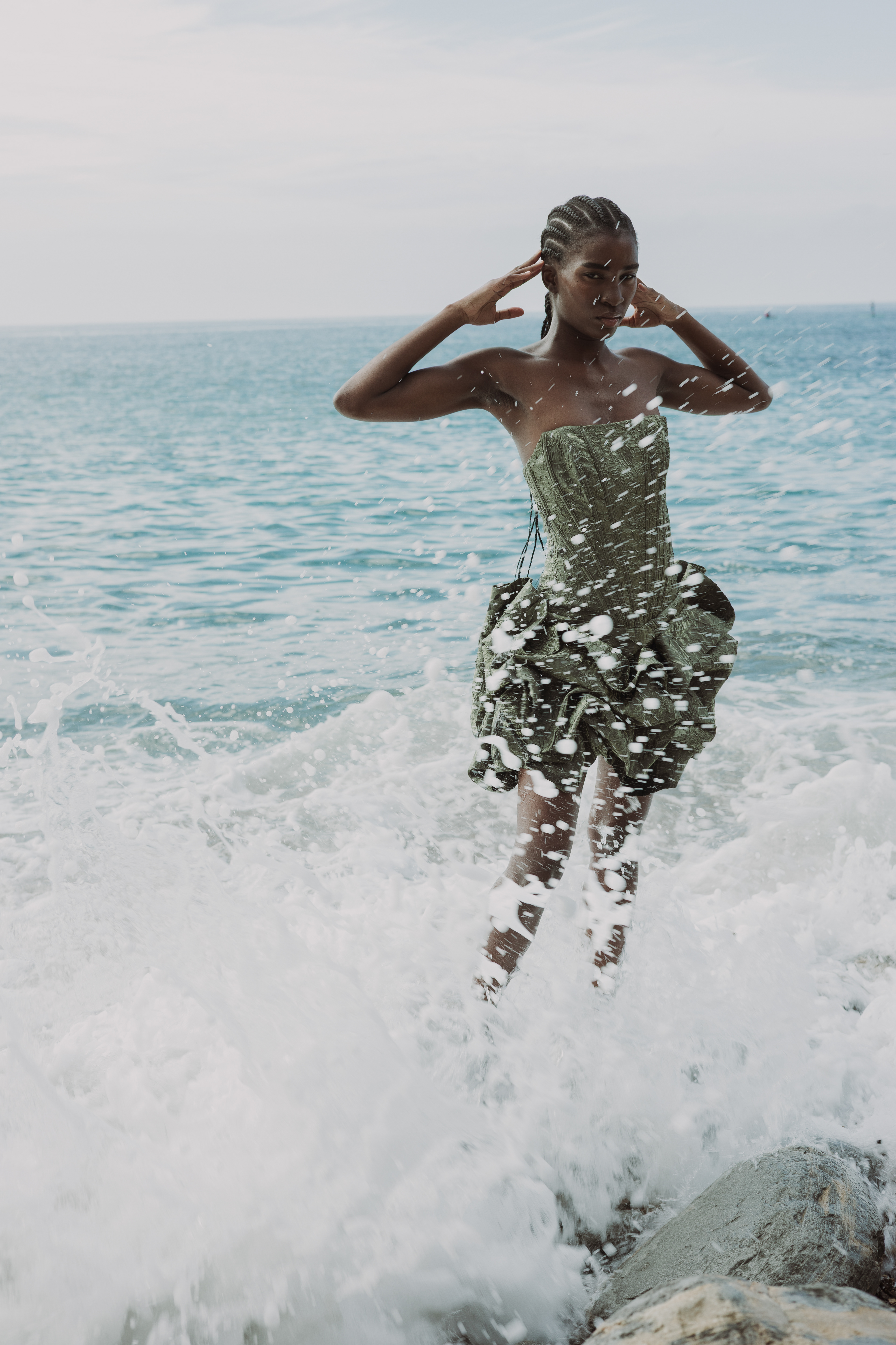 Editorial fashion photograph of a woman wearing a textured green dress, standing amid crashing ocean waves with both hands raised near her head.