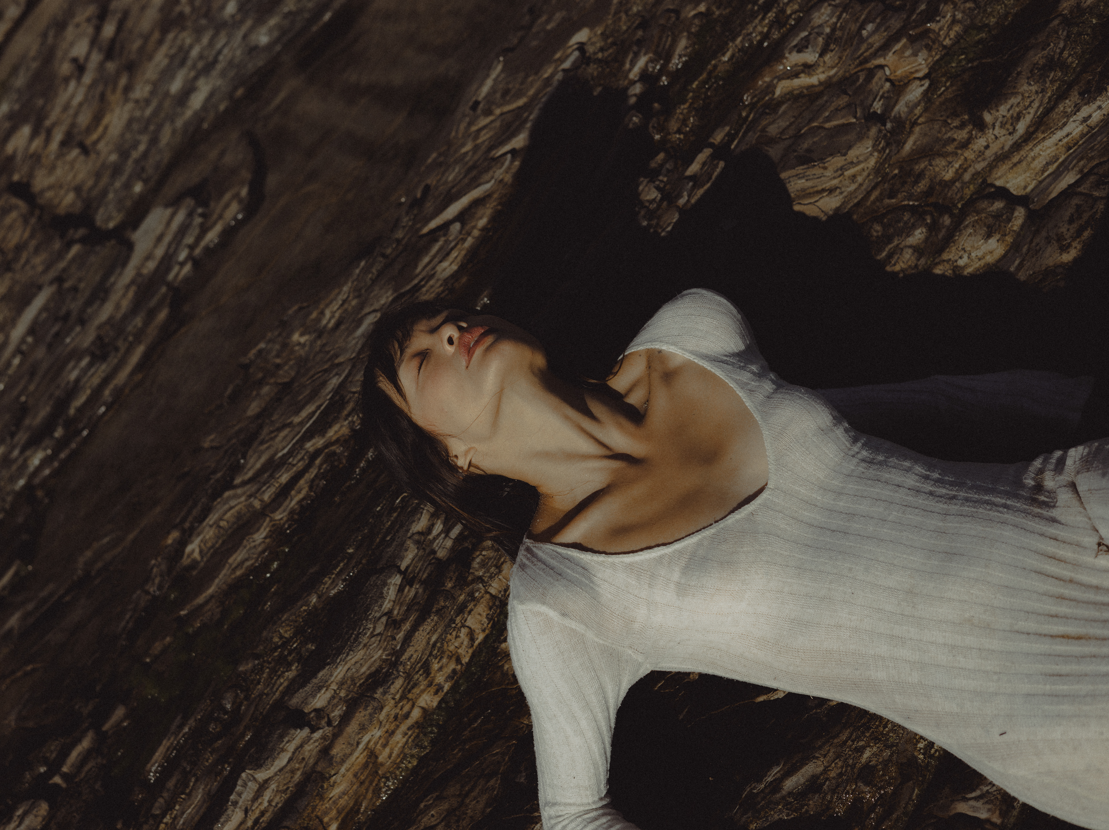 Fashion portrait of a woman in a white ribbed dress reclining against dark, textured coastal rocks, with her head tilted back.