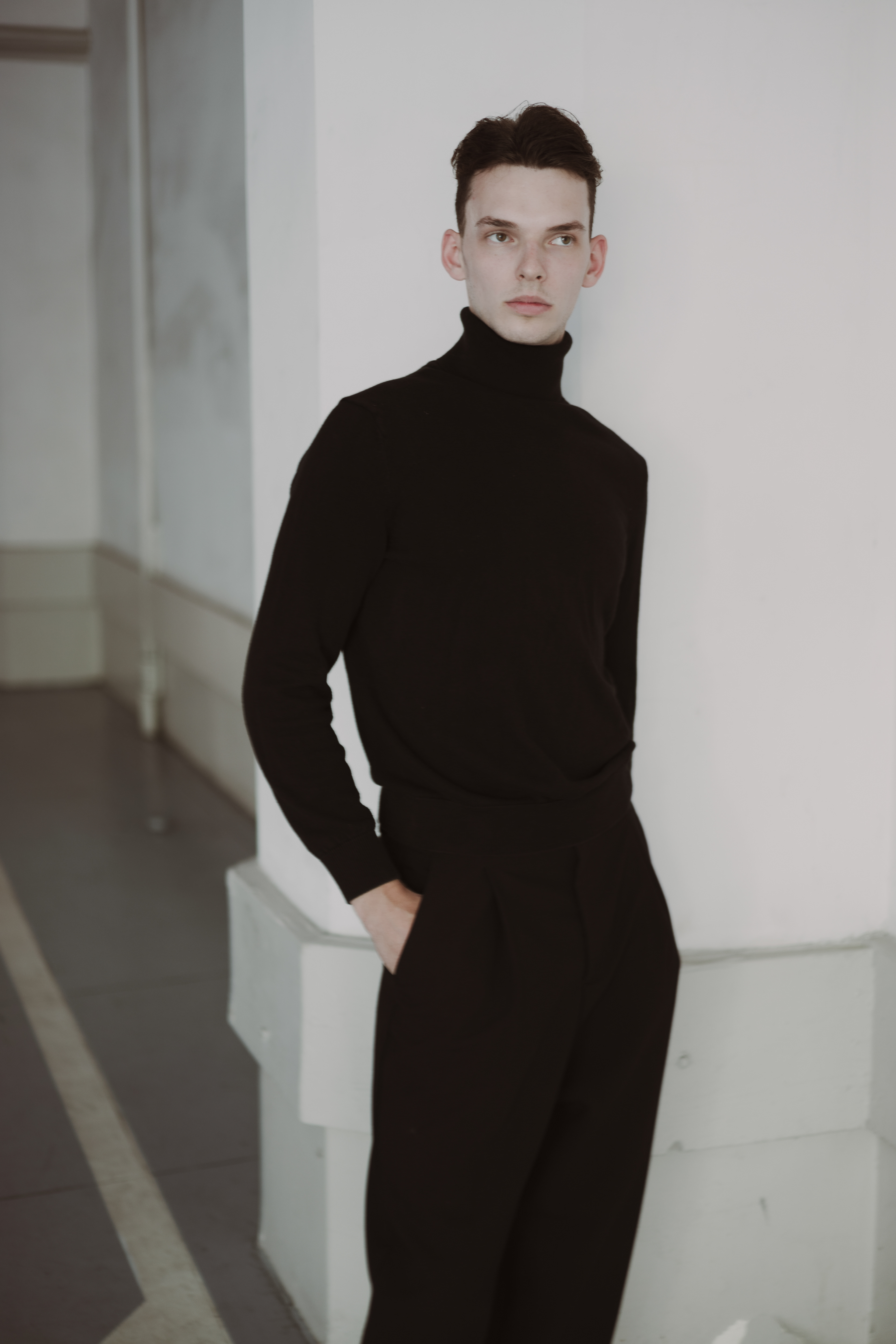Editorial menswear portrait of a male model in a black turtleneck and tailored trousers, standing beside a white architectural column with one hand in his pocket.