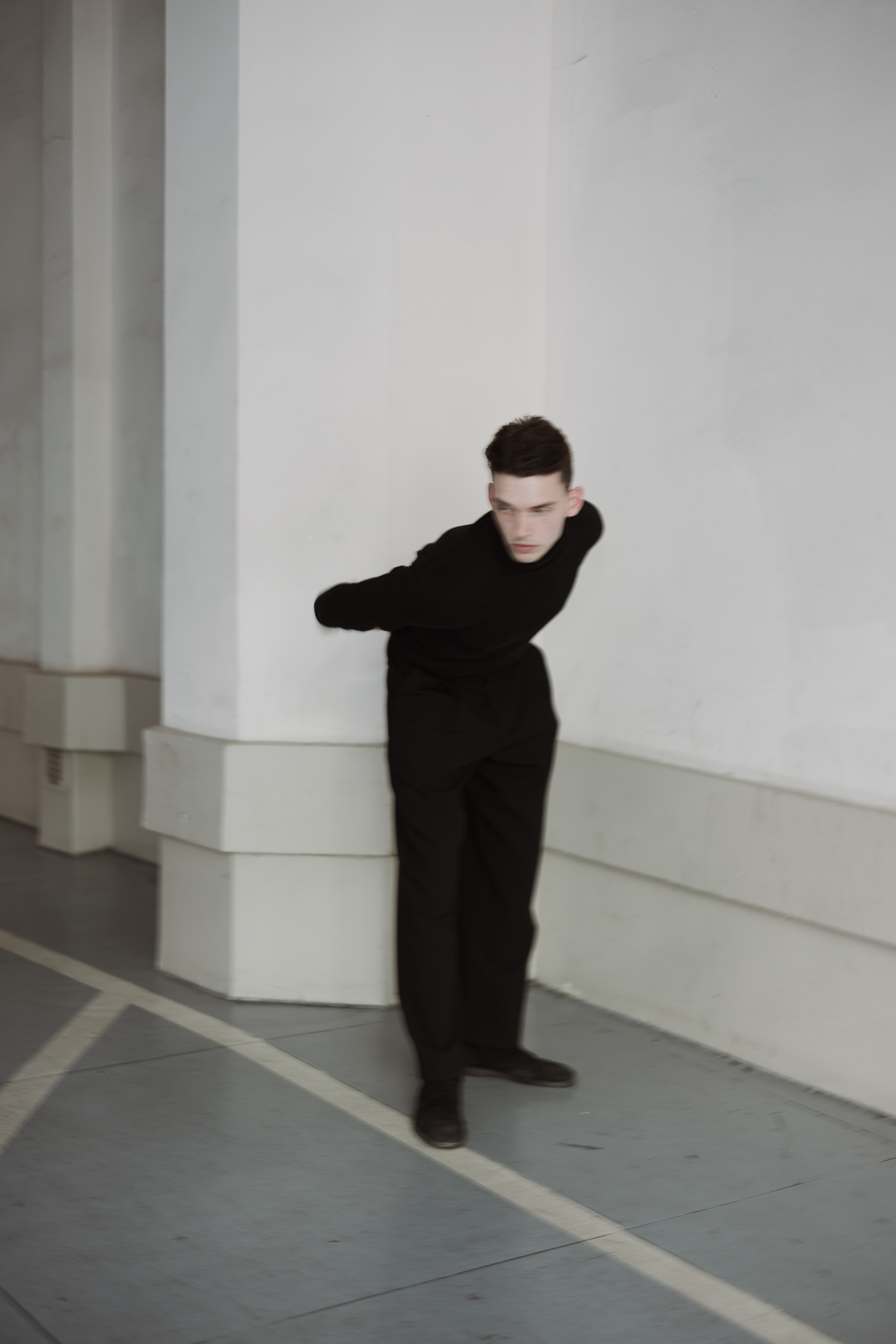 Fashion editorial photograph of a male model dressed in an all-black tailored outfit, leaning forward in a dynamic pose against minimalist white architecture.