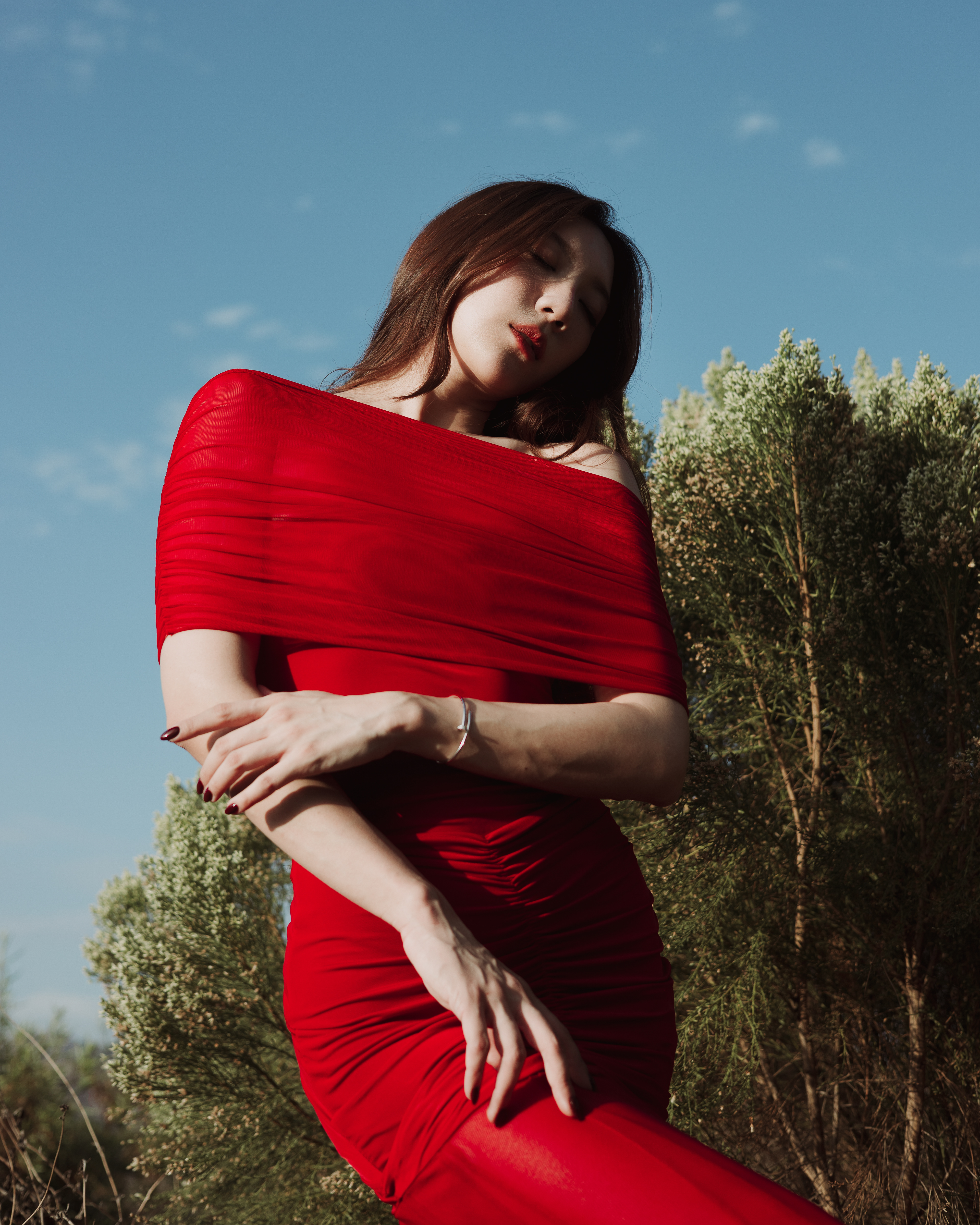 Editorial fashion portrait of a woman wearing a vibrant red off-the-shoulder draped dress, posing with crossed arms against a backdrop of greenery and blue sky.