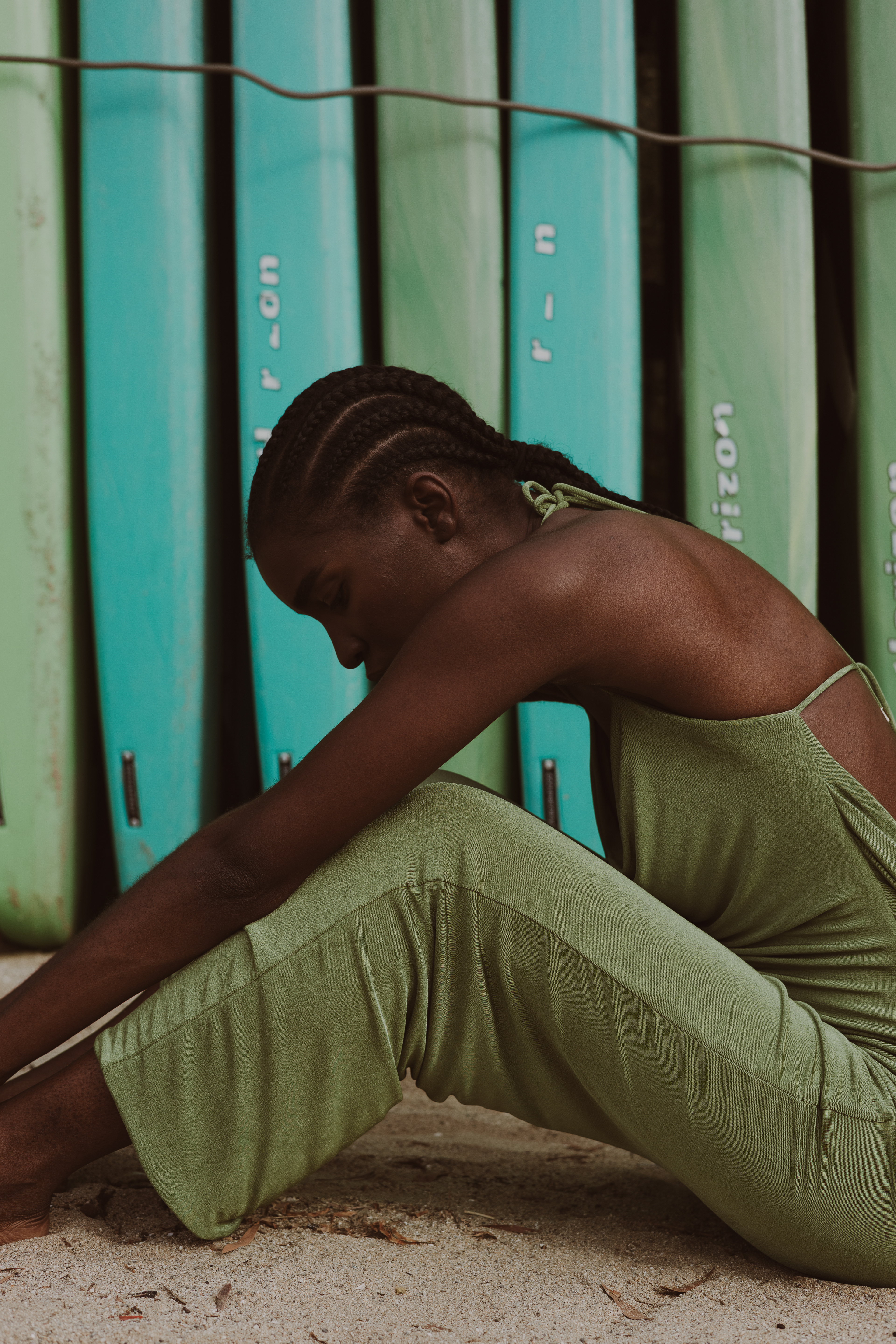 Editorial fashion photograph of a woman in a draped sage-green dress sitting with her head lowered and arms extended in front of turquoise surfboards.