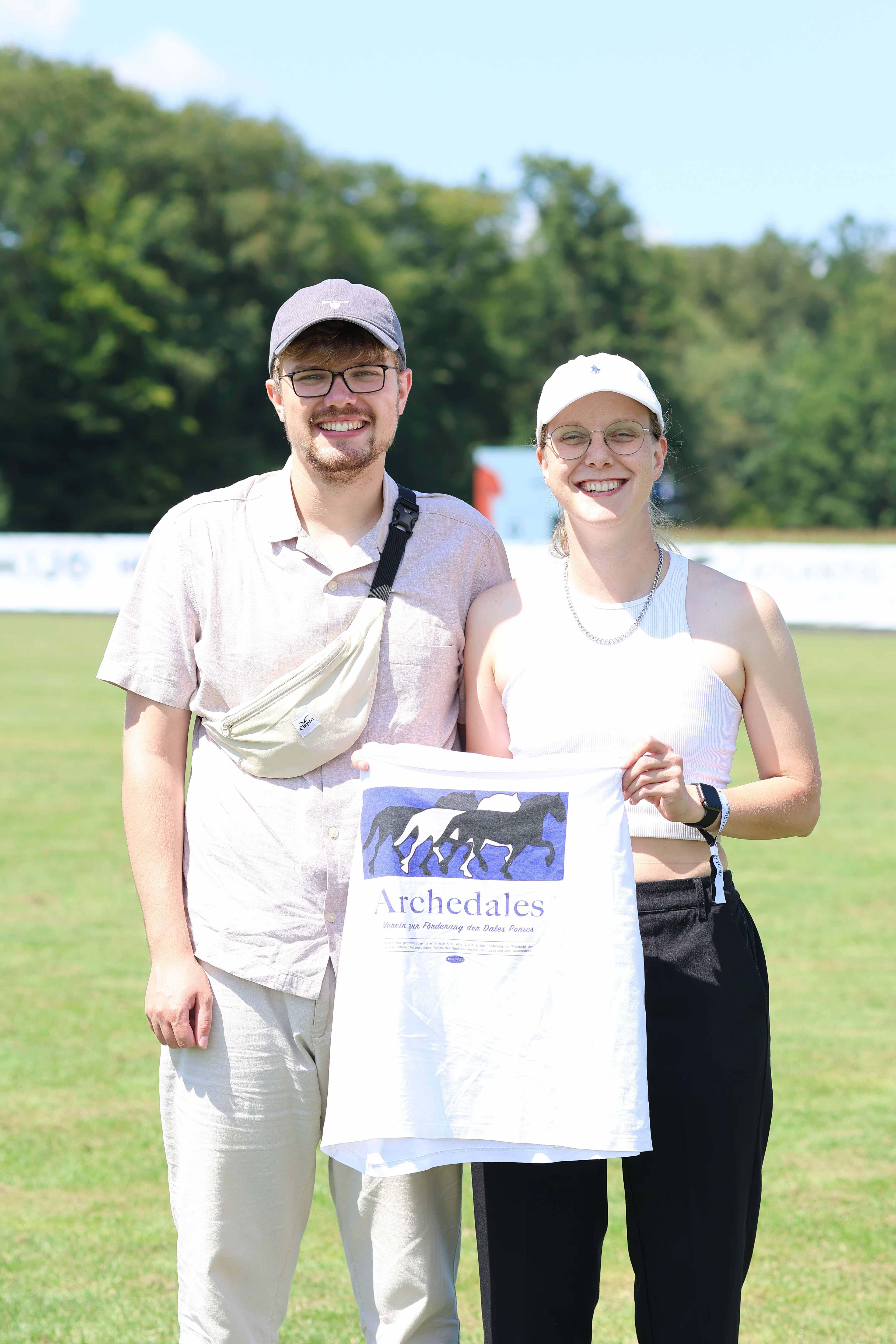 Katharina Borlinghaus und Leon Pielage mit archedales Shirt in der Hand auf einem Turnierplatz.