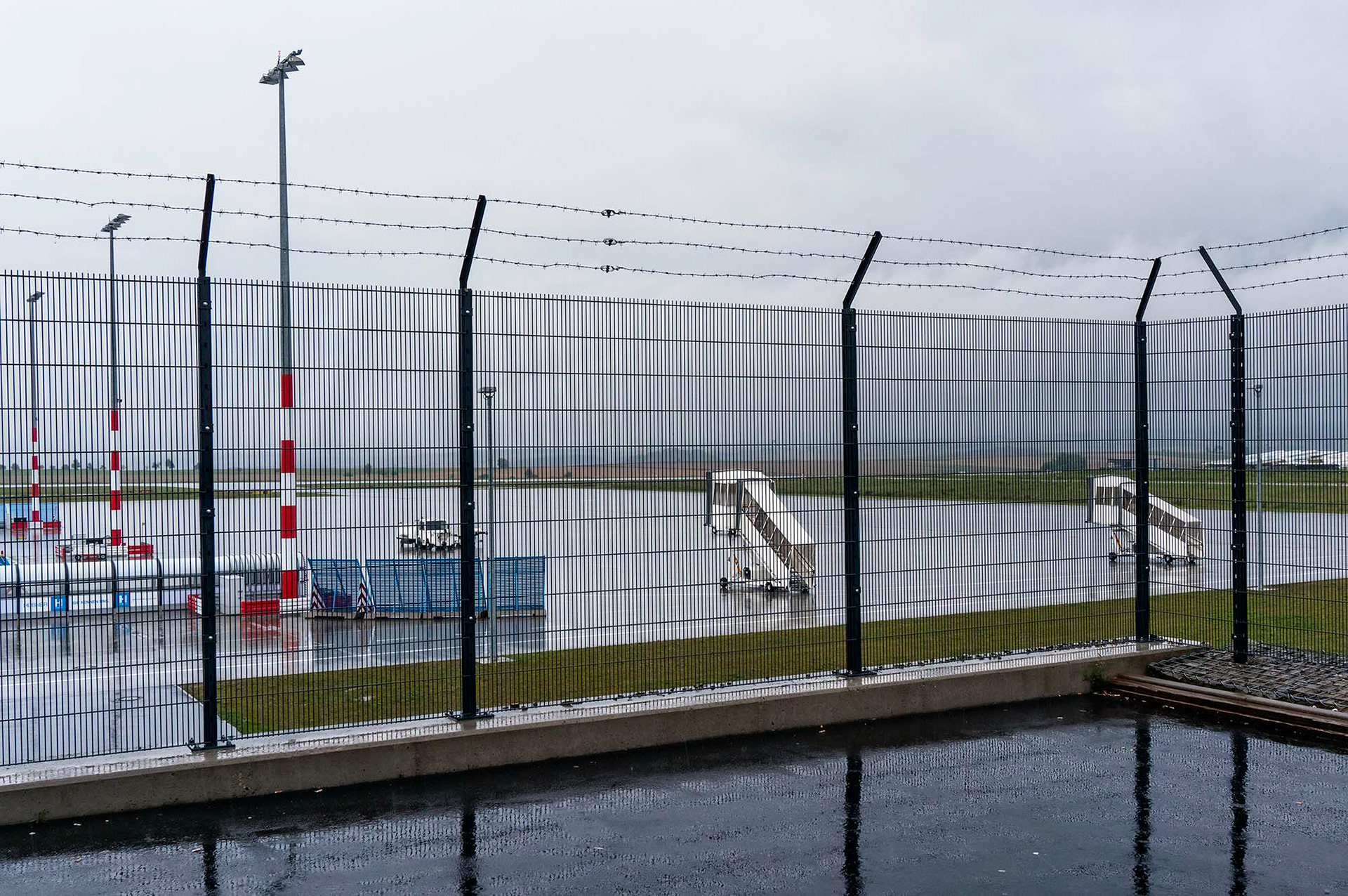Schutzzaun mit Stacheldraht und einem Vorfeld ohne Flugzeuge und Menschen, auf dem zwei einsame Fluggasttreppen im Regen stehen. Flughafen Kassel-Calden. Fotograf Klaus Ohlenschläger - Frankfurt - Bad Homburg – Kronberg