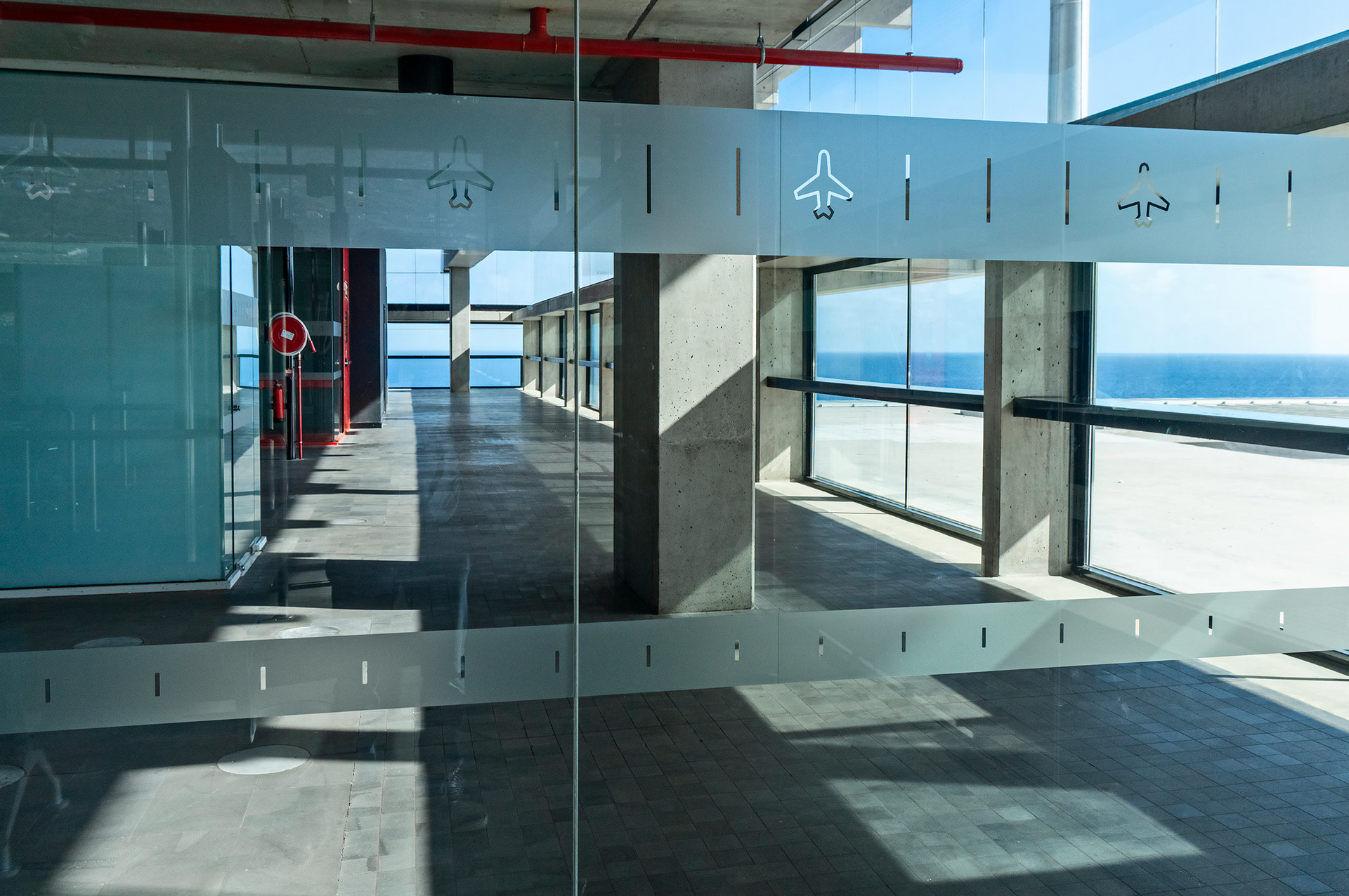 Terminal - Eine Glastür versperrt den Zugang zum leeren Bereich des Gebäudes. Piktogramme mit Flugzeugen. Leere mit Aussicht durch große Fenster auf eine blaue Wasserfläche: Ein Airport ohne Passagiere. Clean, einsam. Keine Verbindung.