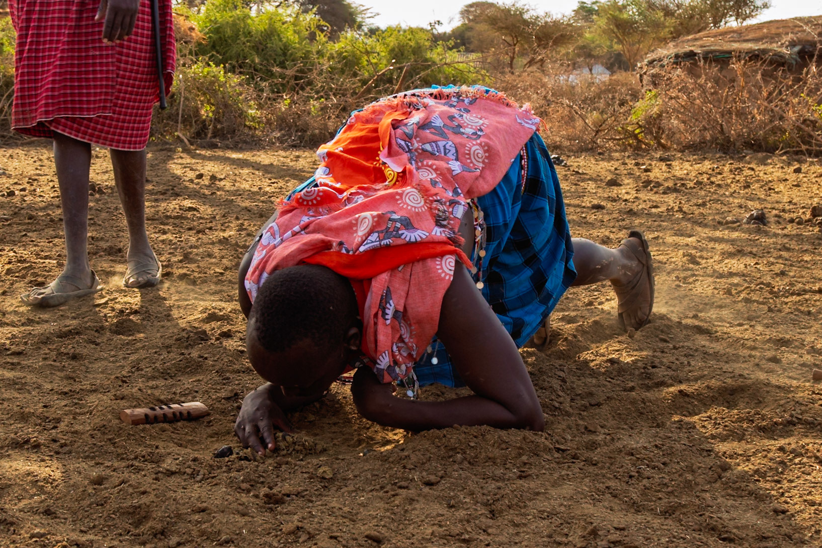 A Maasai man in Kenya kneels on the ground, possibly in a ritual or ceremony, while another person stands nearby.