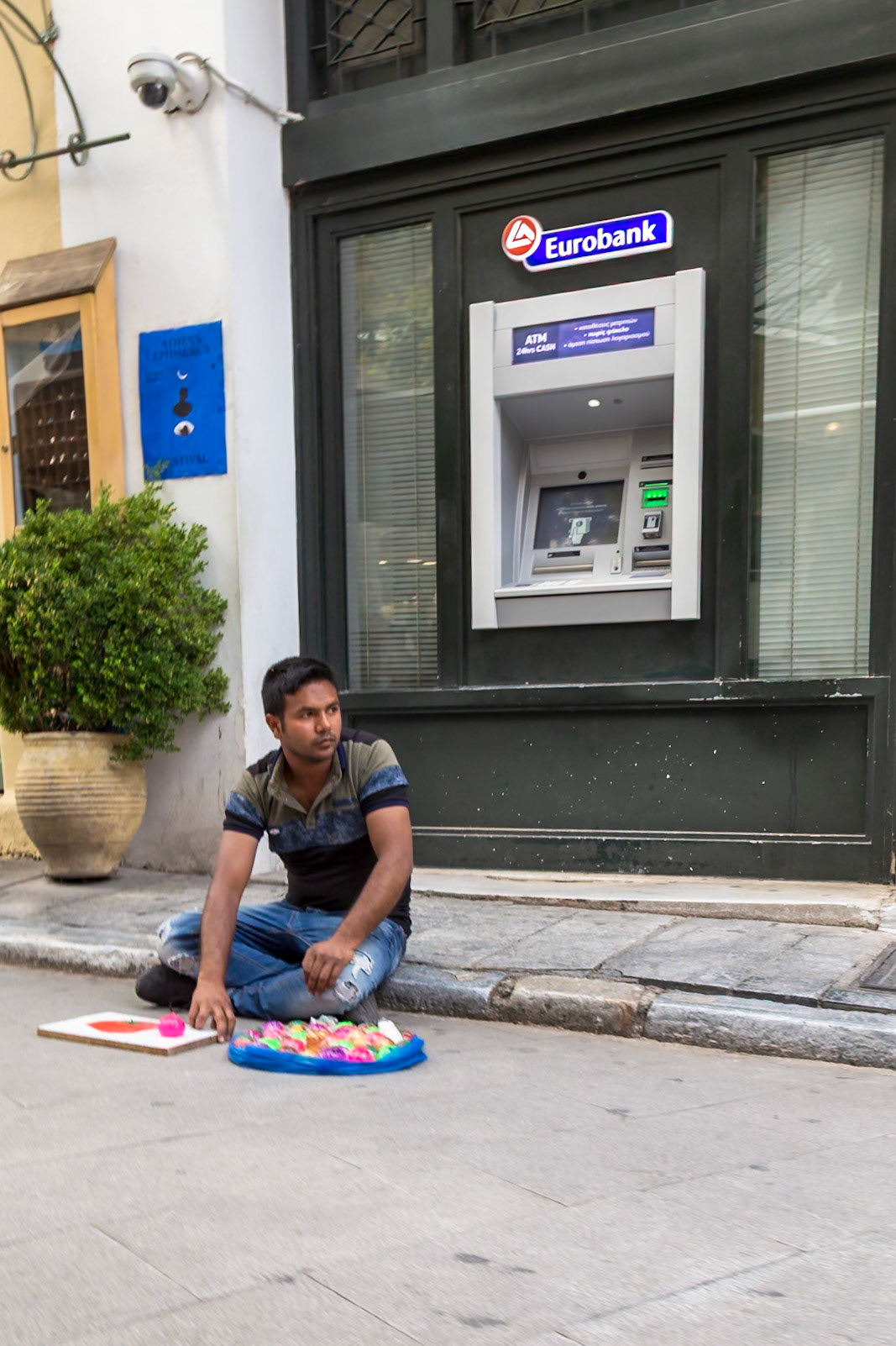 Athens, Greece - May 23rd 2018: A street vendor sits near a Eurobank ATM, displaying colorful toys for sale to passersby to earn a living.