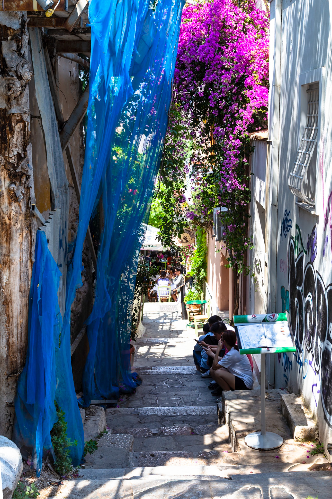 Athens, Greece - May 23rd 2018: People sit on steps in a narrow street, looking at their phones, while a restaurant server stands nearby.