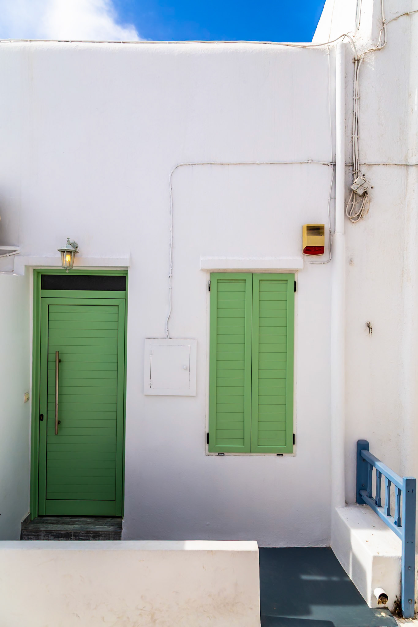Mykonos, Greece - May 22nd 2018: A building's exterior is shown, featuring a green door and window shutters against a white wall, showcasing Cycladic architecture.