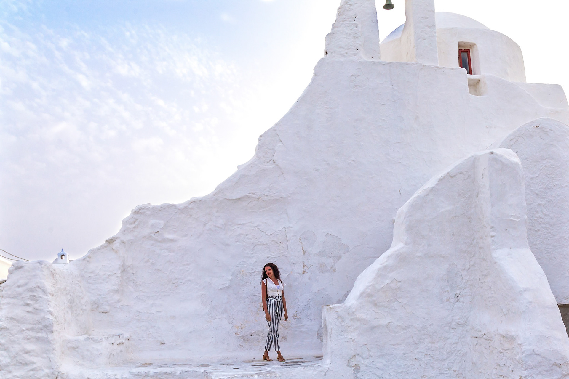 Mykonos, Greece - May 23rd 2018: A woman in striped pants poses by the Paraportiani Church, a whitewashed architectural landmark.
