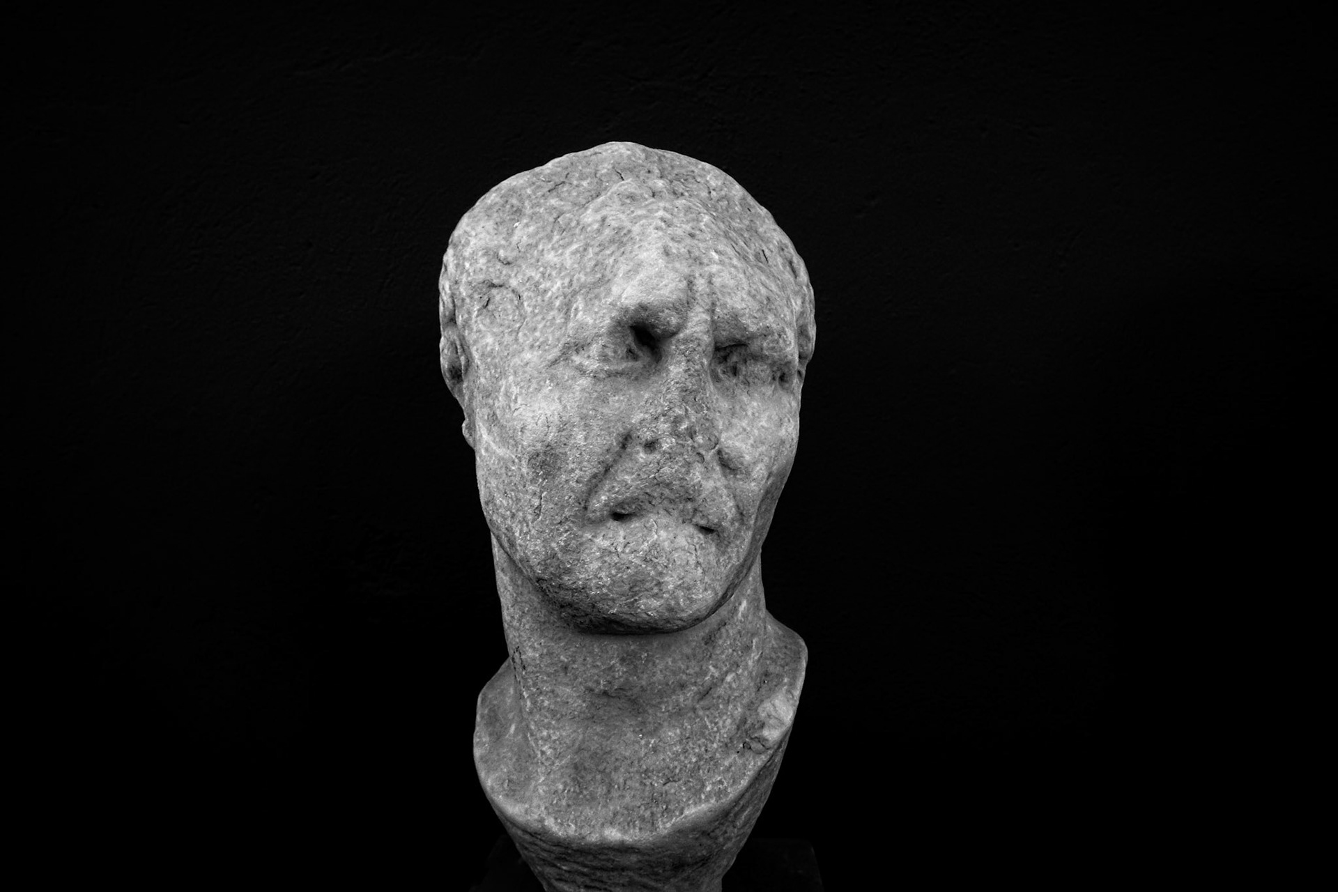 Delos, Greece - May 22nd 2018: A weathered, stone bust of a man stands against a black background, showcasing ancient Greek sculpture and artistry.