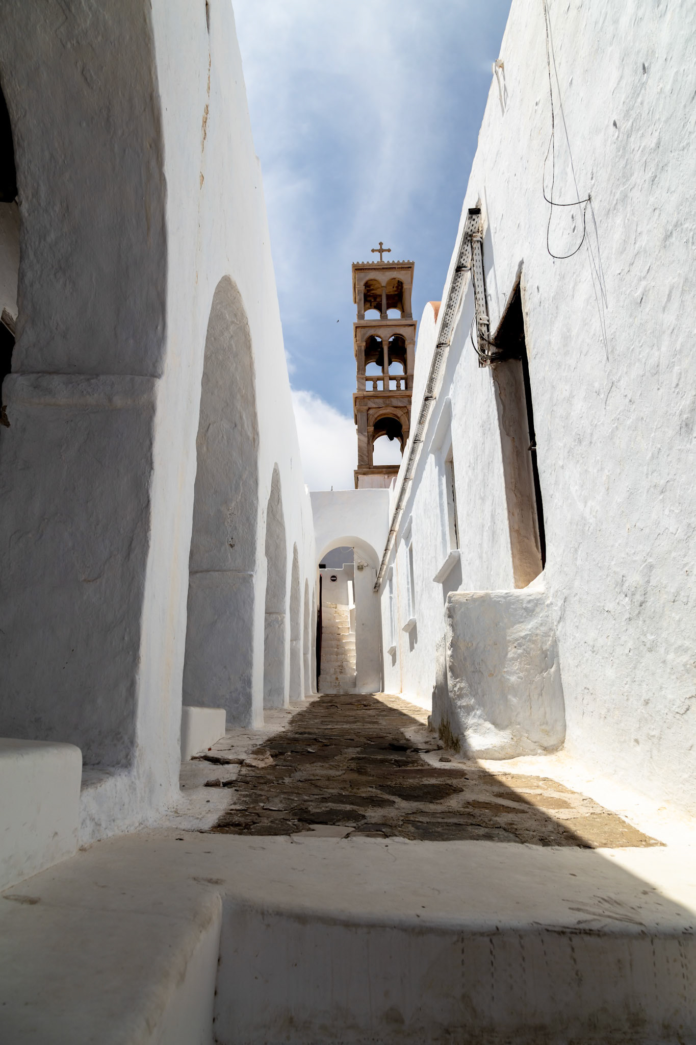 Mykonos, Greece - May 22nd 2018: A narrow, stone-paved street winds between whitewashed buildings, leading to a distant church tower under a blue sky.