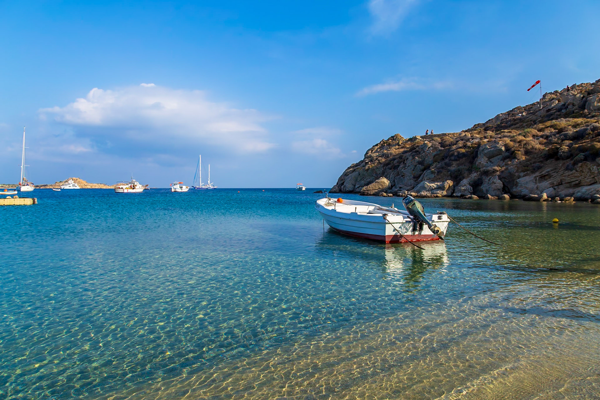 Psarou Beach, Mykonos, Greece - May 24th 2018: A small boat is moored in the clear water, with people visible on the hill in the background.