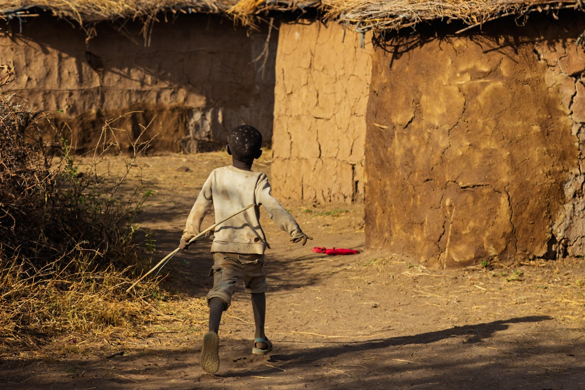 A Maasai child walks through a Kenyan village, stick in hand, past traditional mud huts.