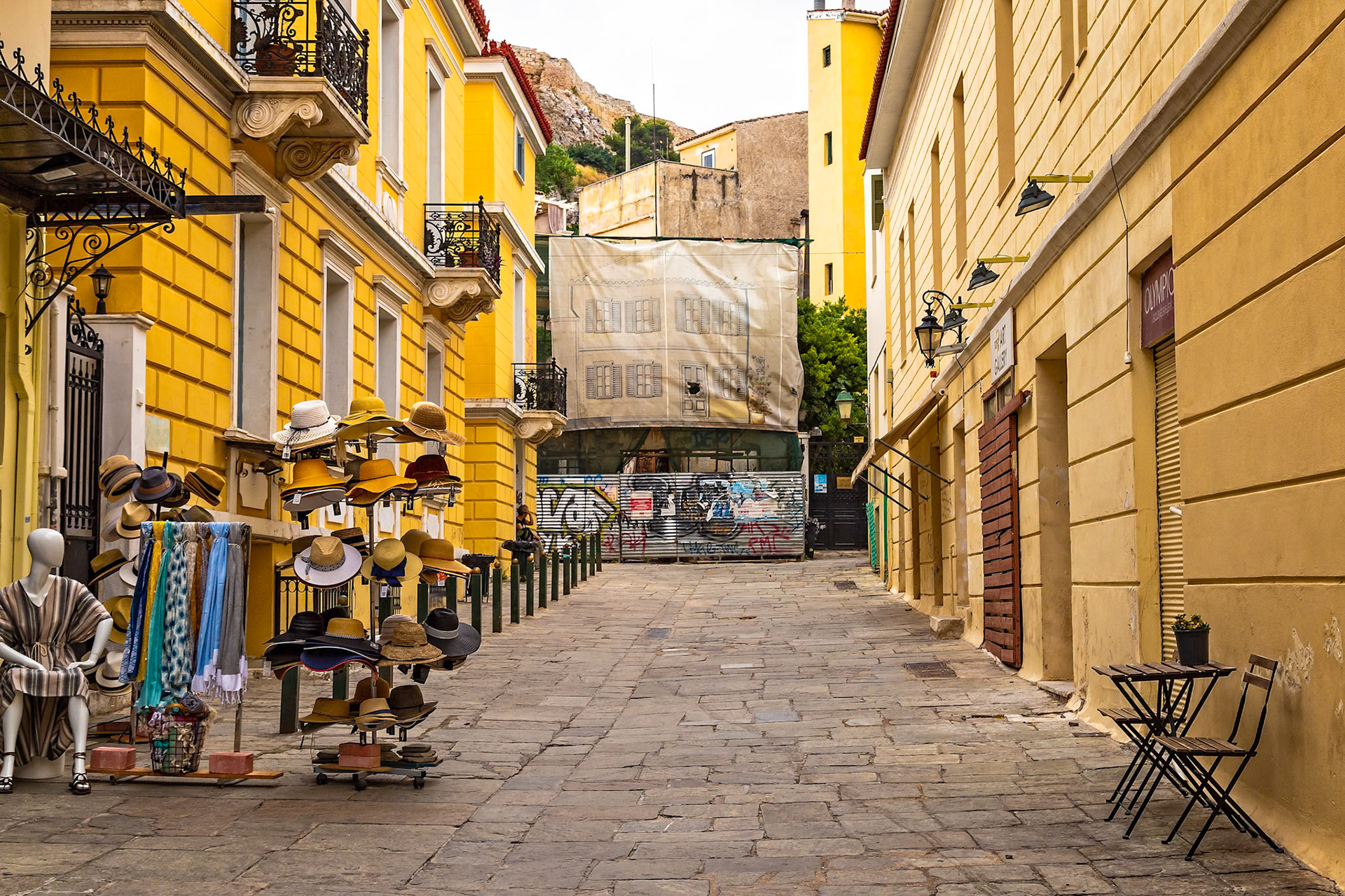 Athens, Greece - May 23rd 2018: A street vendor displays hats and scarves for sale on a narrow street lined with yellow buildings, attracting tourists.
