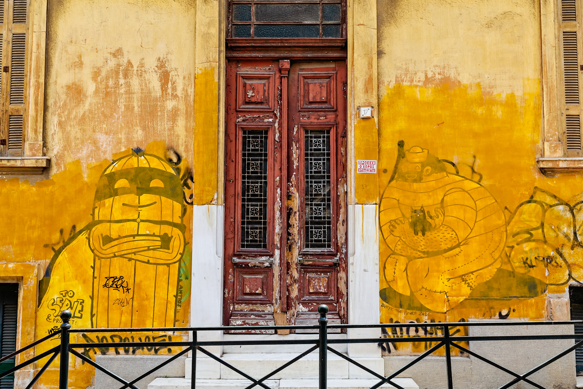 Athens, Greece - May 23rd 2018: A building with graffiti art on the yellow walls, flanking a weathered red door. The art depicts cartoonish figures.