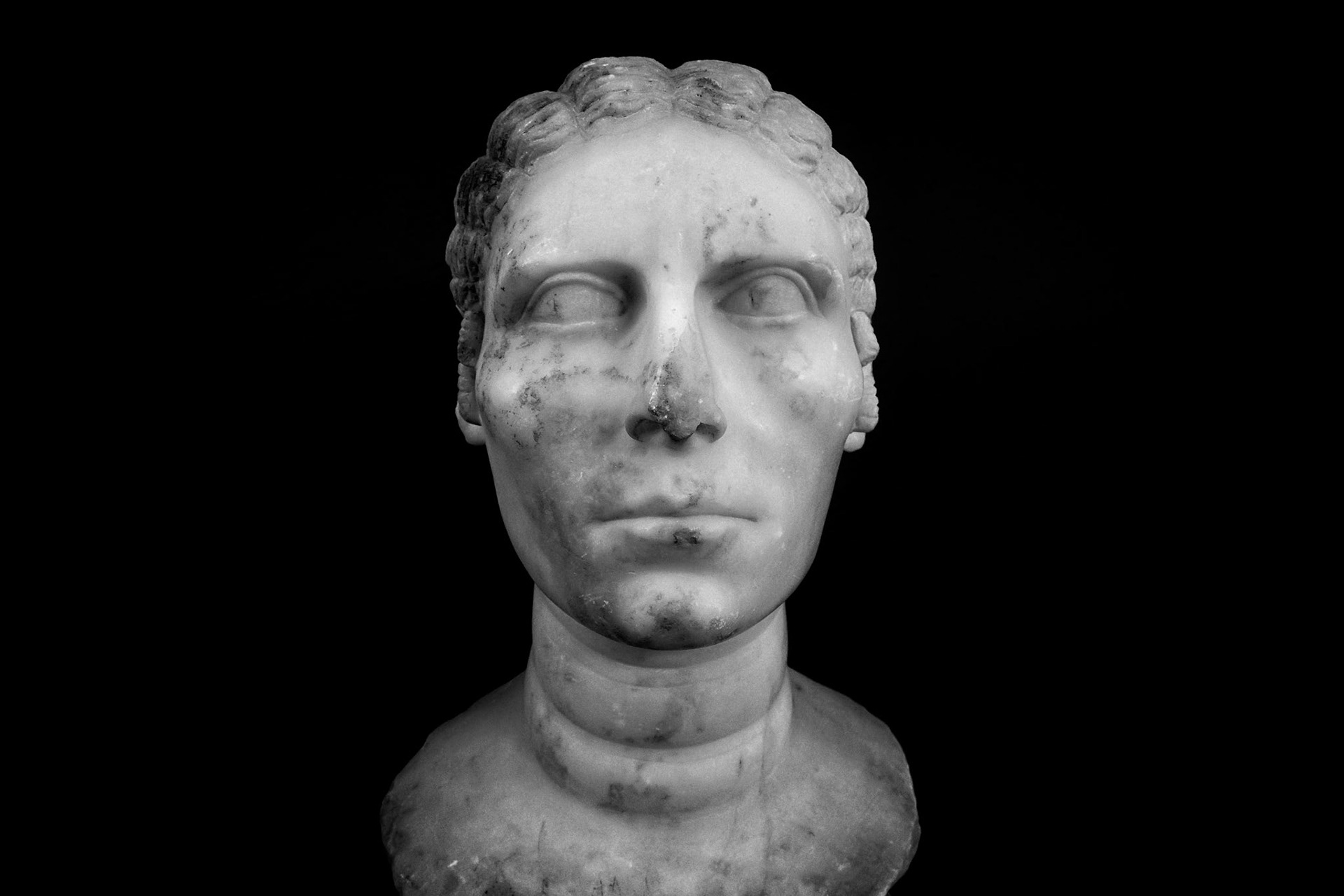 Delos, Greece - May 22nd 2018: A marble bust of a woman with her hair pulled back, likely a depiction of a goddess or noblewoman, stands against a black backdrop.